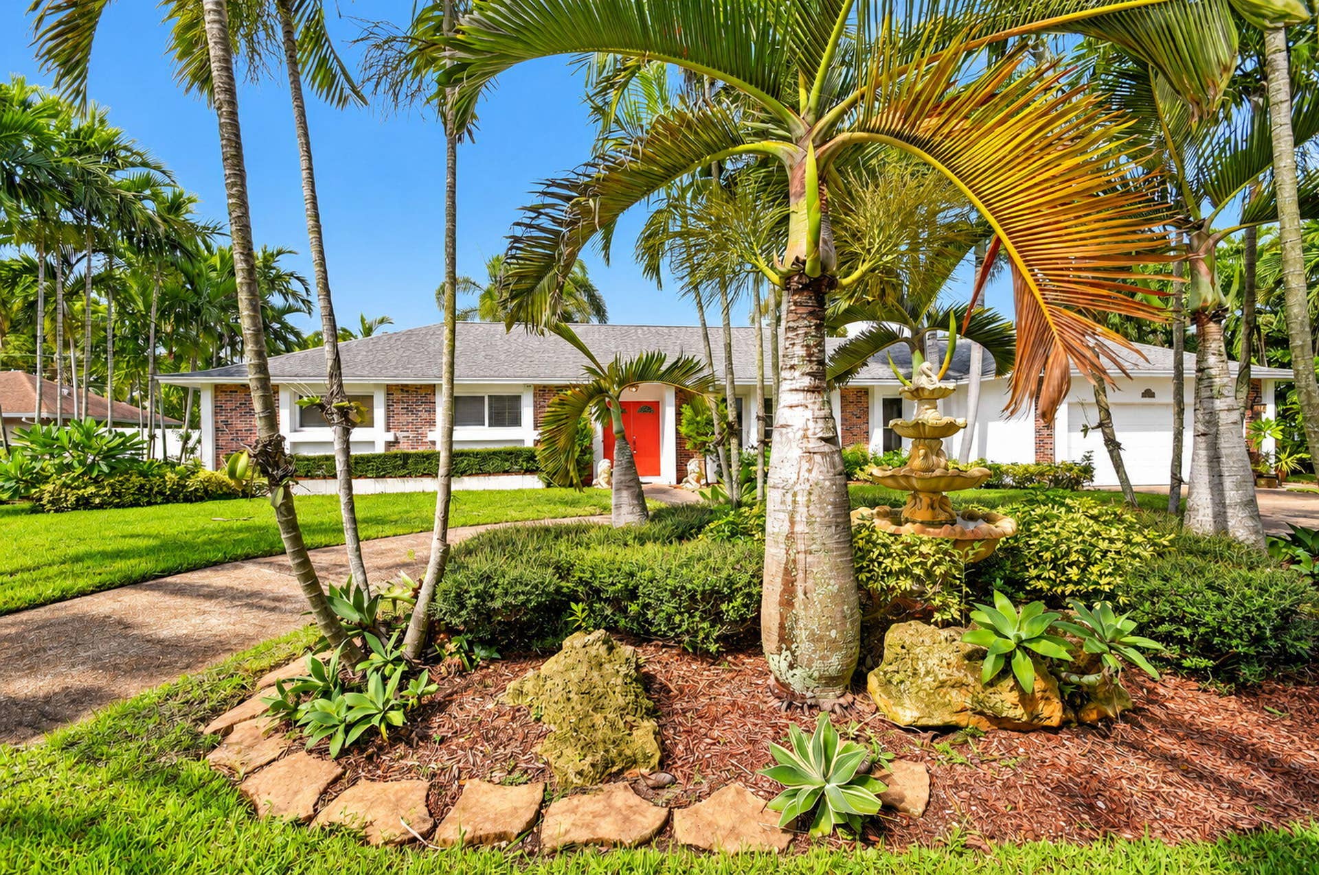 Expansive front yard with a tiered water feature, mature palm trees, diverse plantings, and a textured pathway