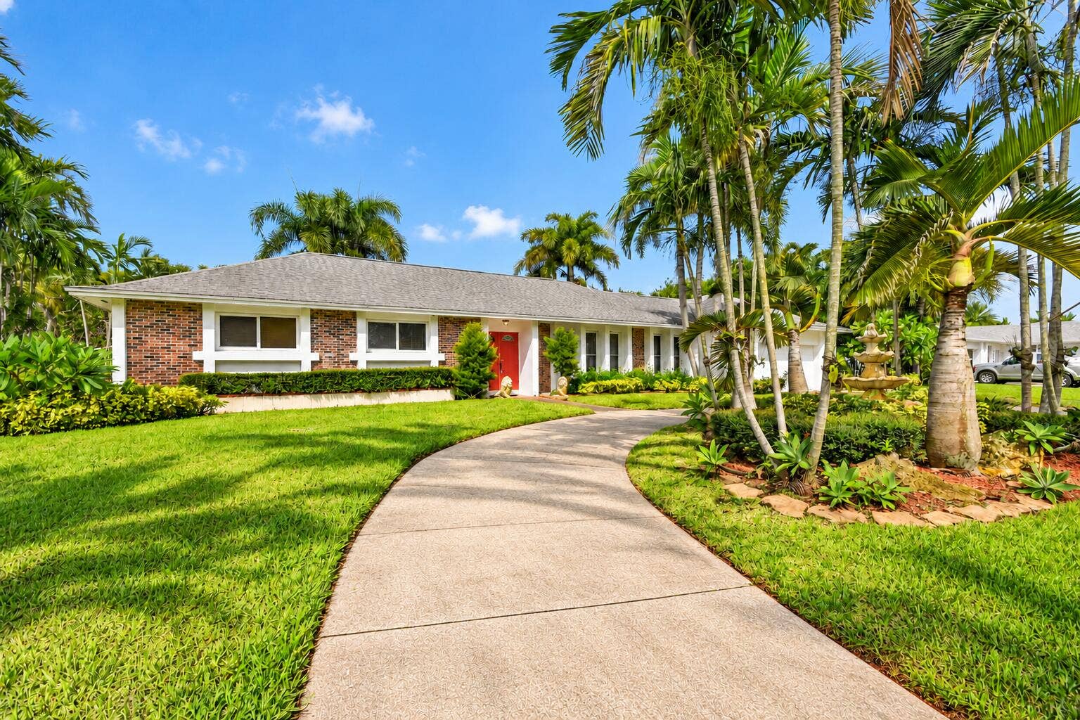 Brick facade residence featuring a red entry door, a curvilinear concrete driveway, mature palm trees, and a multi-tiered fountain