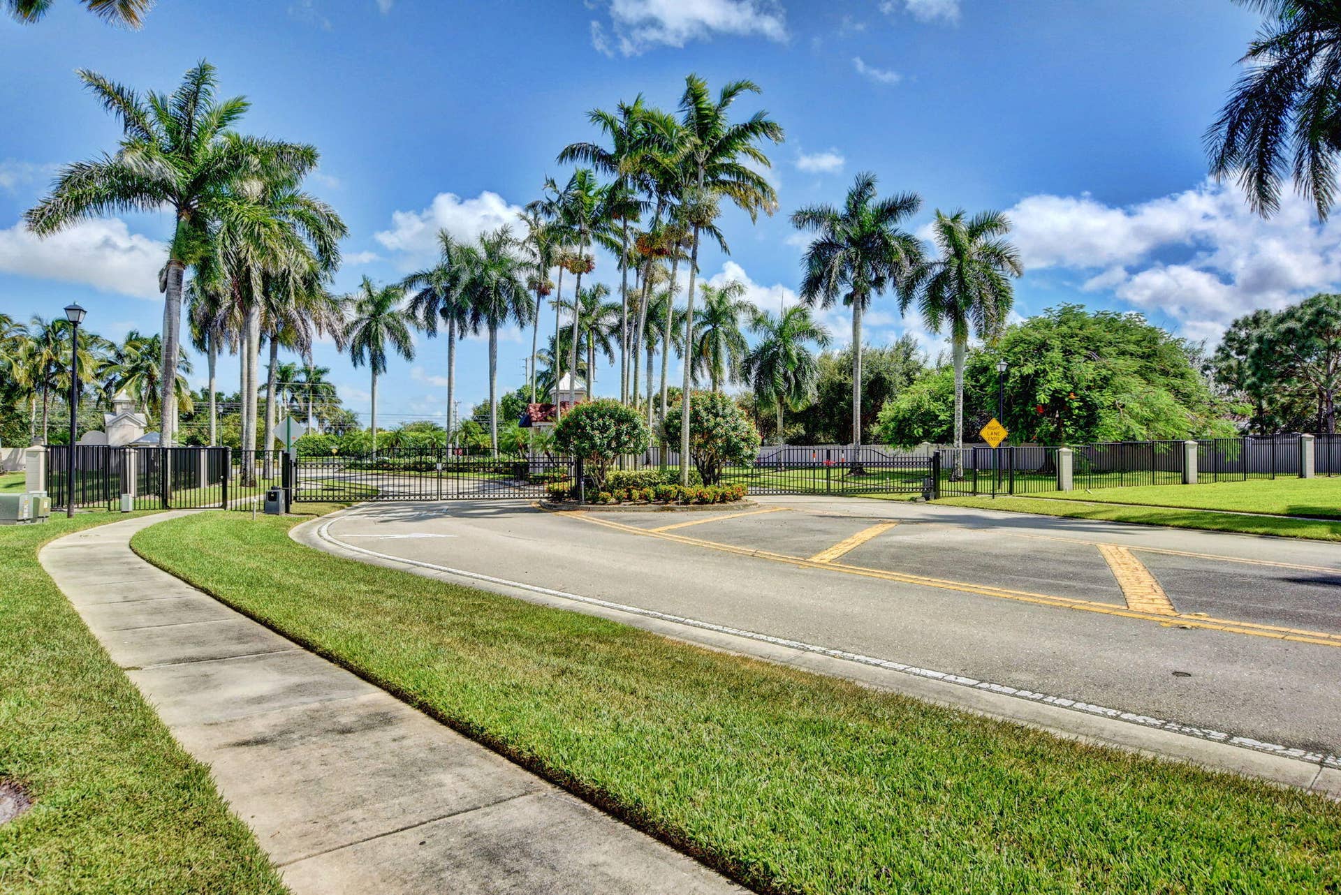 6610 Aston Lane, Boynton Beach, FL 33437 Gated entry featuring a paved road, concrete sidewalk, abundant palm trees, and lush green landscaping