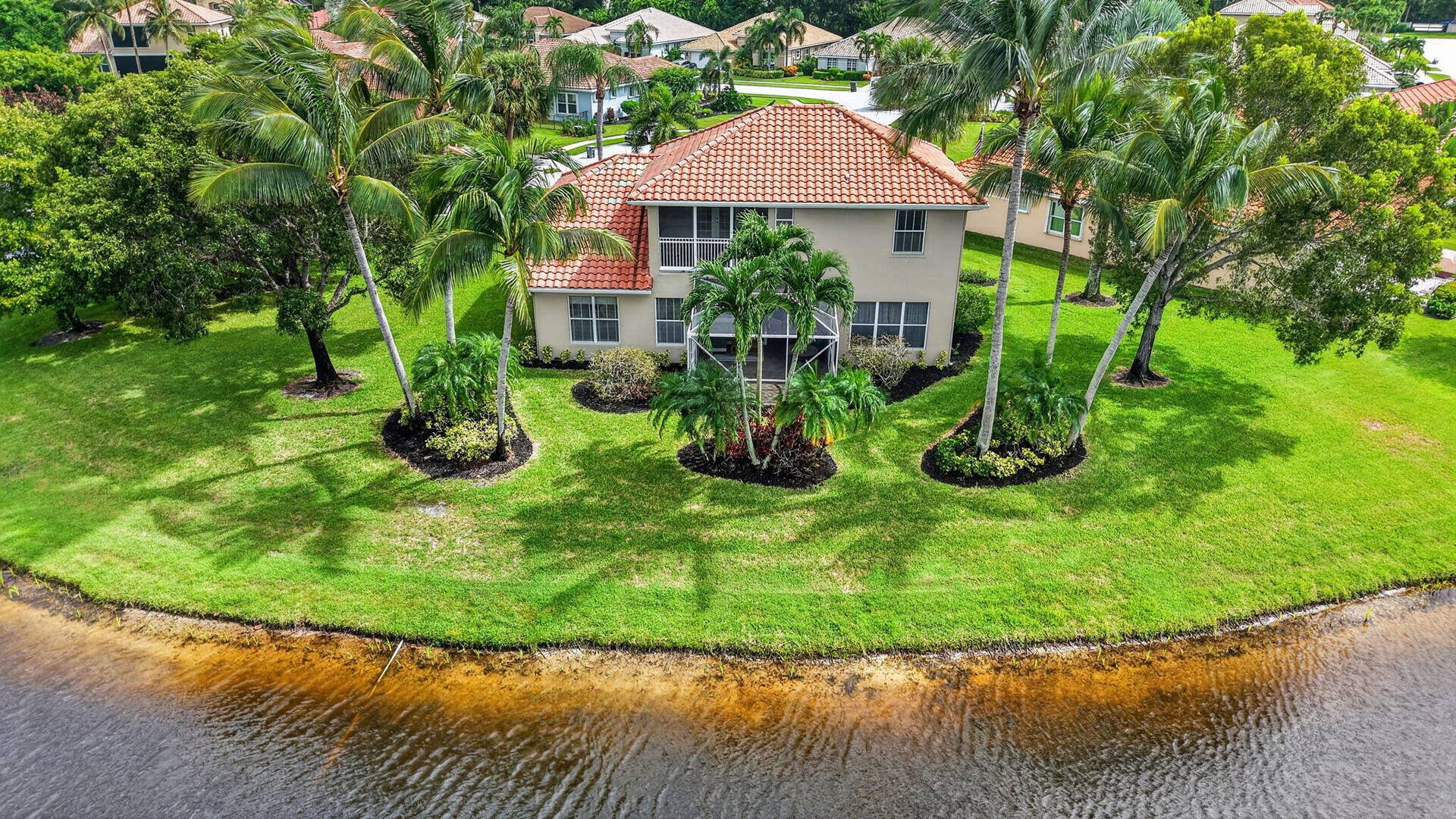 6610 Aston Lane, Boynton Beach, FL 33437 Expansive waterfront property featuring a two-story residence with a red tile roof, screened porch, and verdant landscaping with mature palm trees