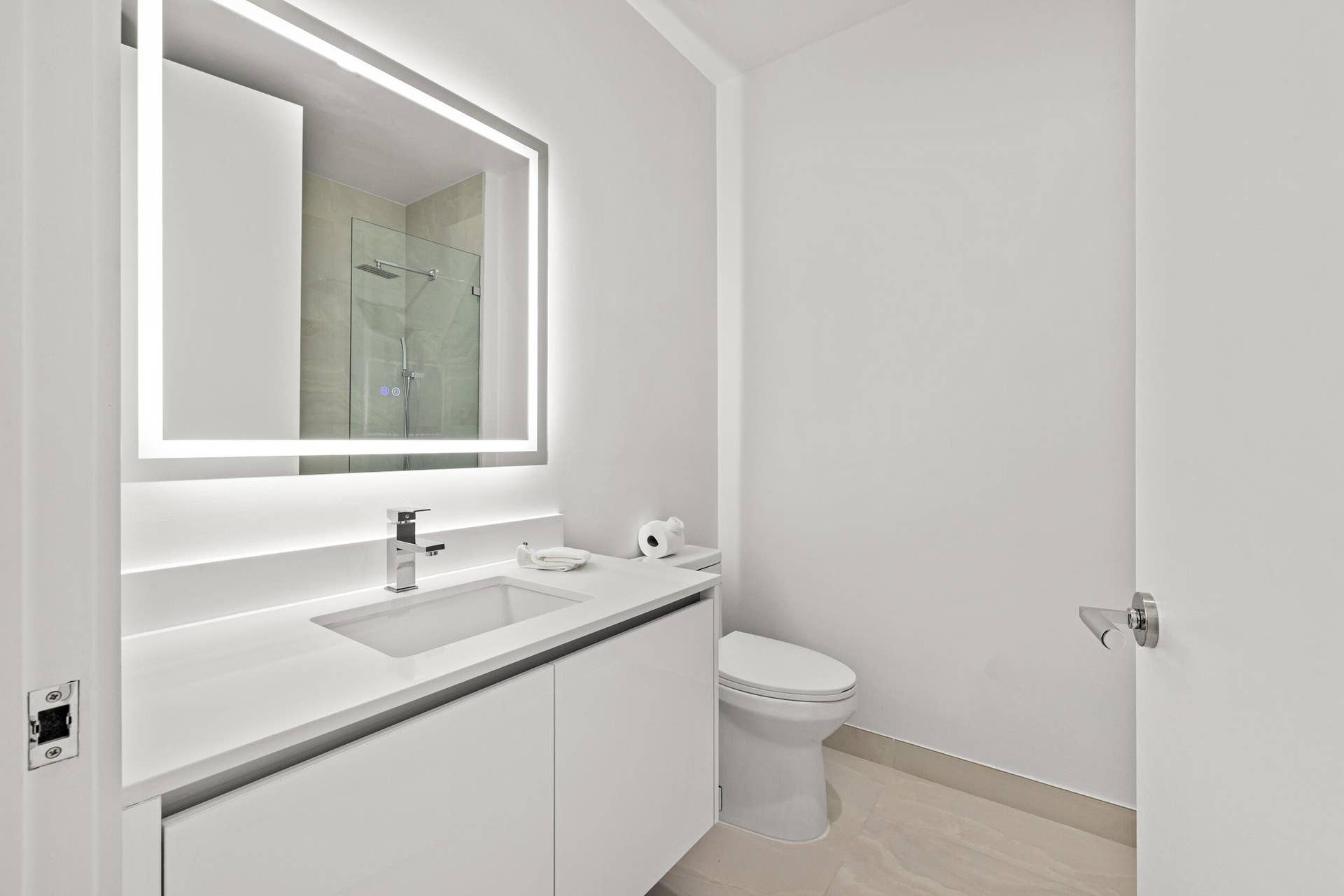 220 Lake Shore Drive, #704, Lake Park, FL 33403 Backlit vanity mirror, single basin sink, white integrated countertop, white cabinetry, and a walk-in shower with glass enclosure