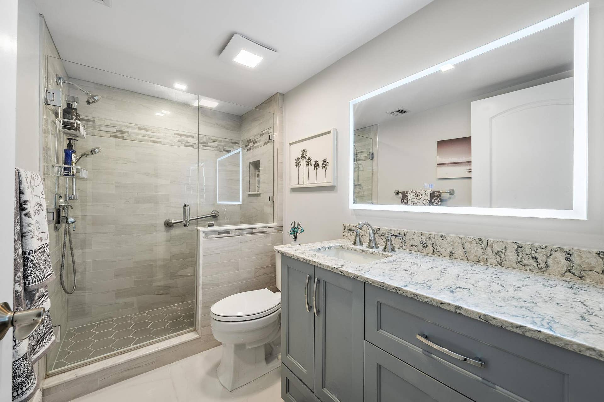 Spacious bathroom featuring a glass-enclosed shower, built-in shower niche, hexagon tile shower floor, single vanity with light-colored countertop, and an illuminated mirror