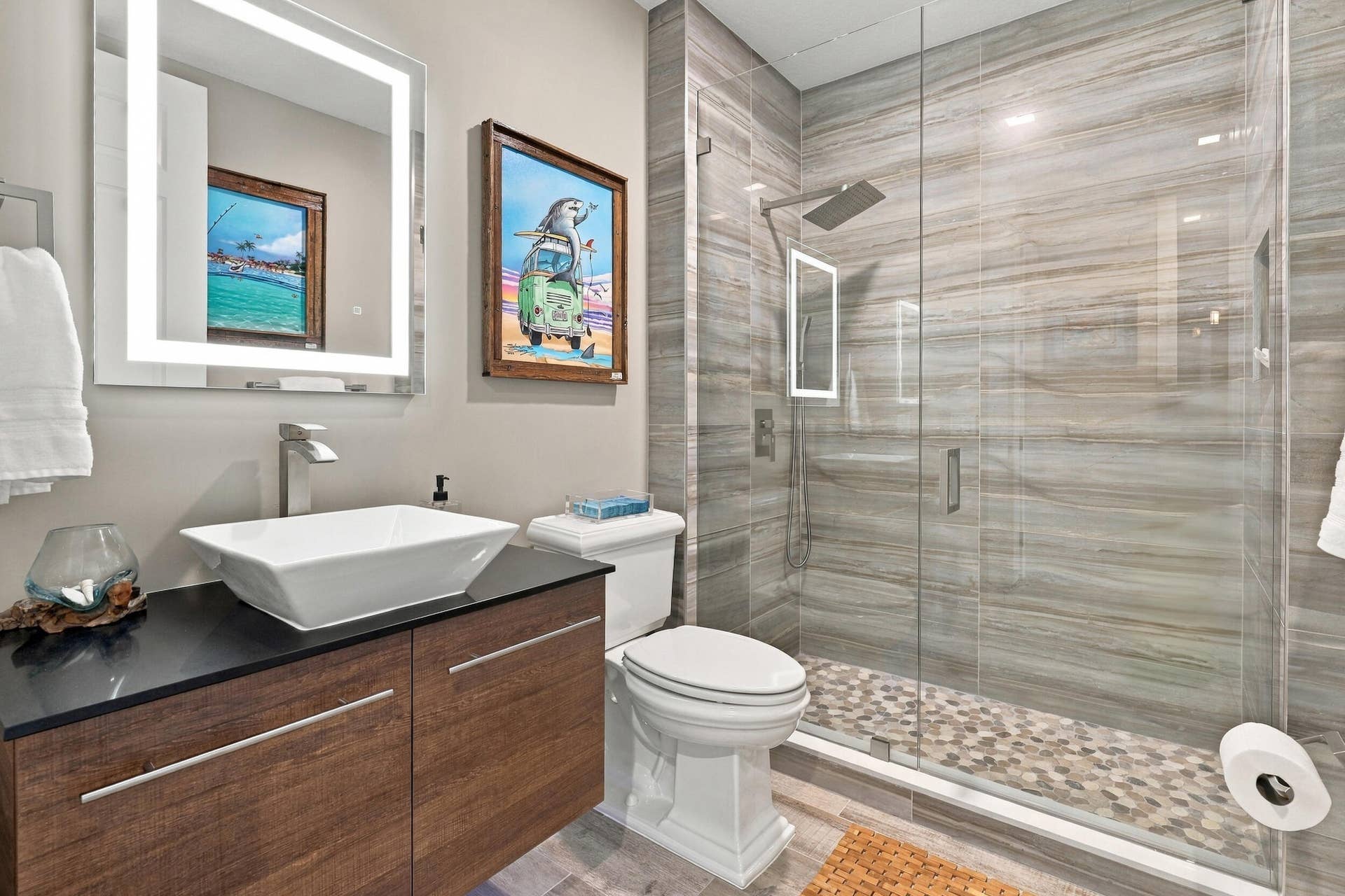 Guest bath features a floating vanity with under lighting and vessel sink, frameless, walk-in glass shower with Rain Showerhead, pebble-like flooring and built-in shower niche, and an LED, de-fog mirror.