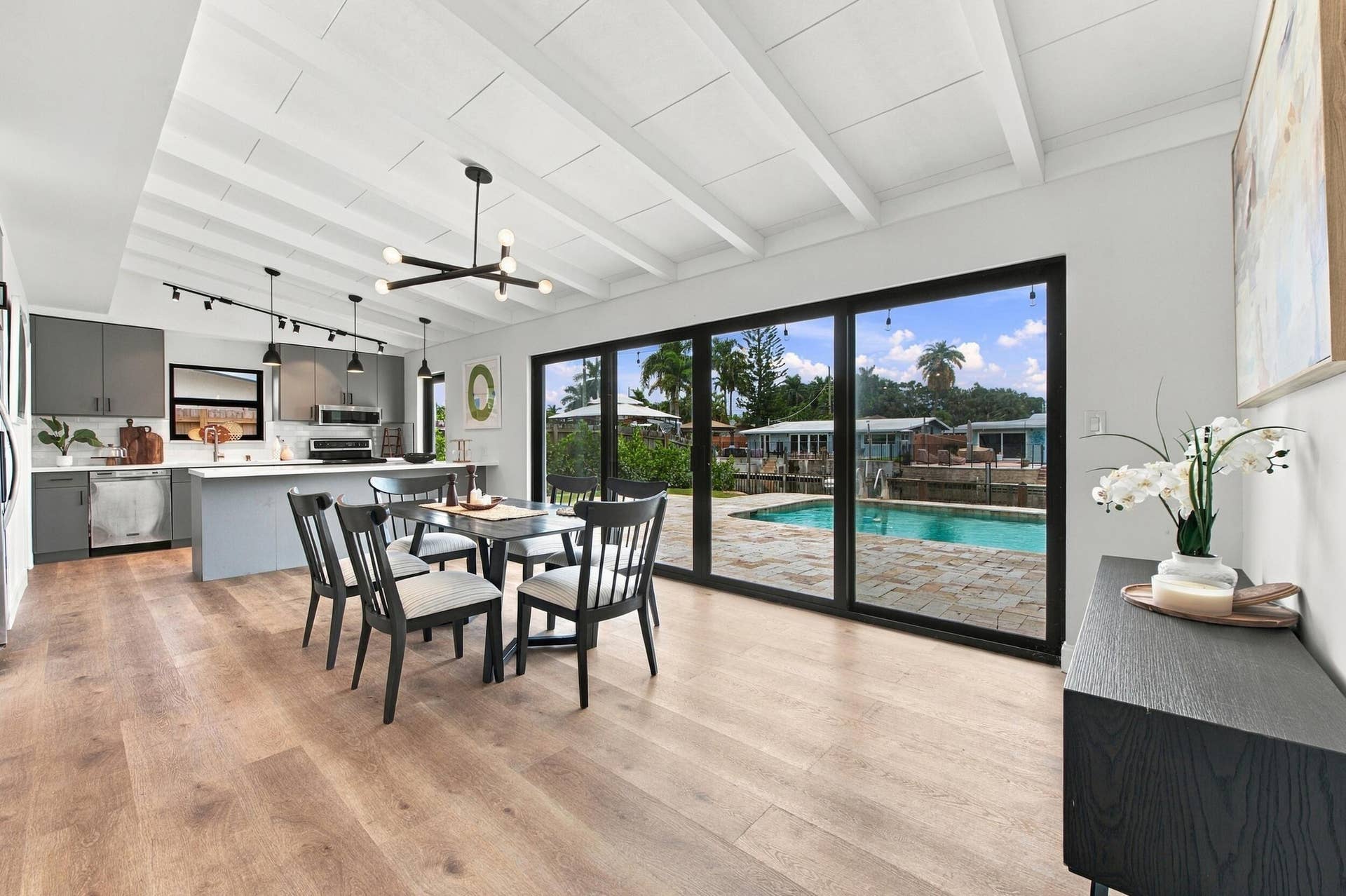 2536 GULFSTREAM Lane, Fort Lauderdale, FL 33312 Open-concept living space with wood-finish flooring throughout, featuring a dining area with seating for four, a fully equipped kitchen with stainless steel appliances and gray cabinetry, and large black-framed sliding glass doors opening to an outdoor entertaining area with resort-style pool