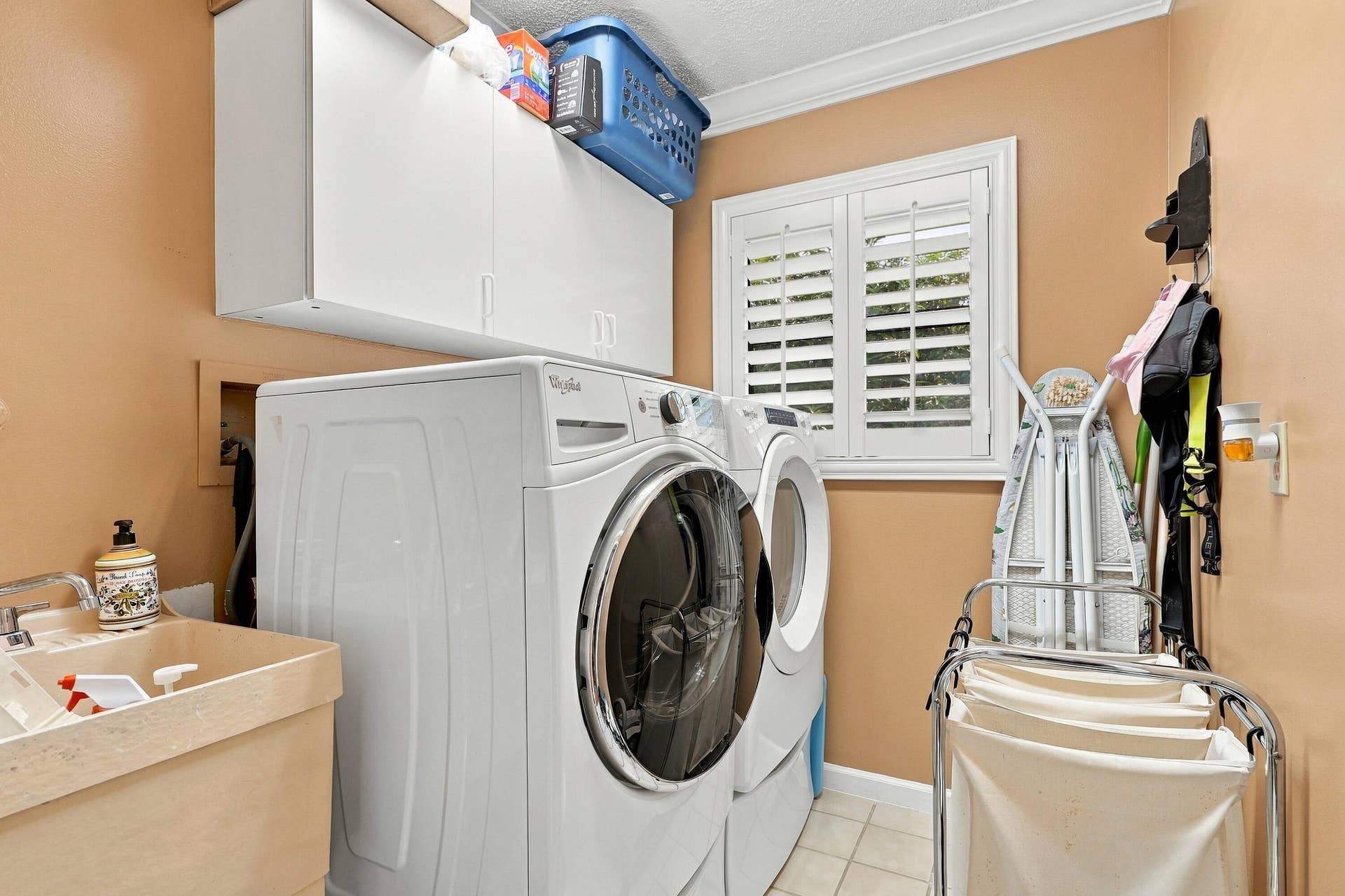 Integrated laundry area featuring white upper cabinetry, a utility sink, and a window with plantation shutters