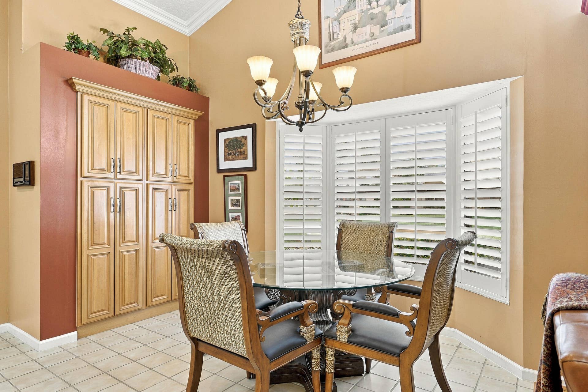 Bay window with white plantation shutters, vaulted ceiling with crown molding, overhead chandelier, light-toned built-in cabinetry, and tile flooring
