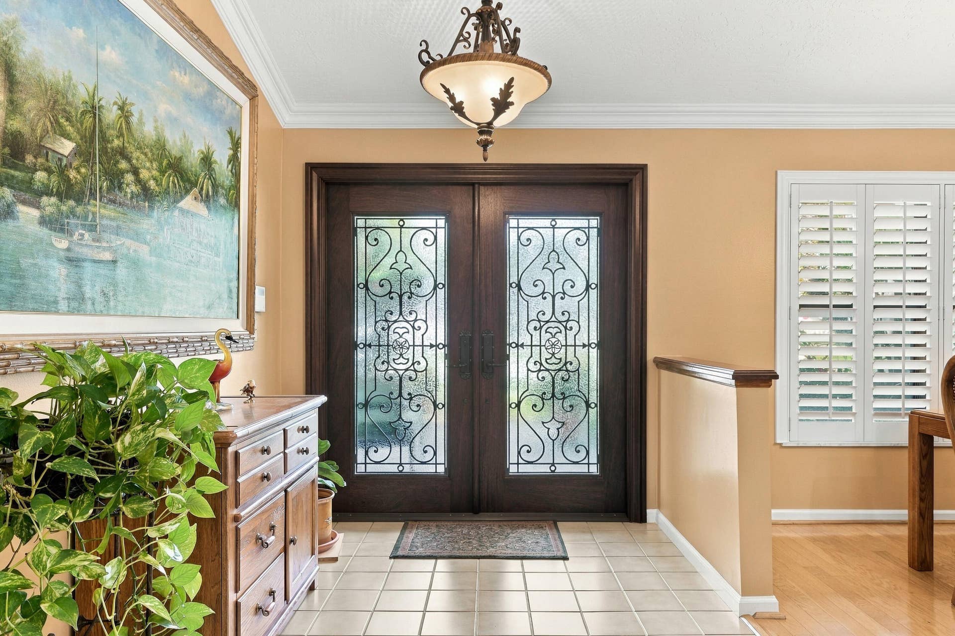 Double entry doors with decorative wrought iron inserts, tiled flooring, elegant ceiling-mounted light fixture, crown molding, and plantation shutters