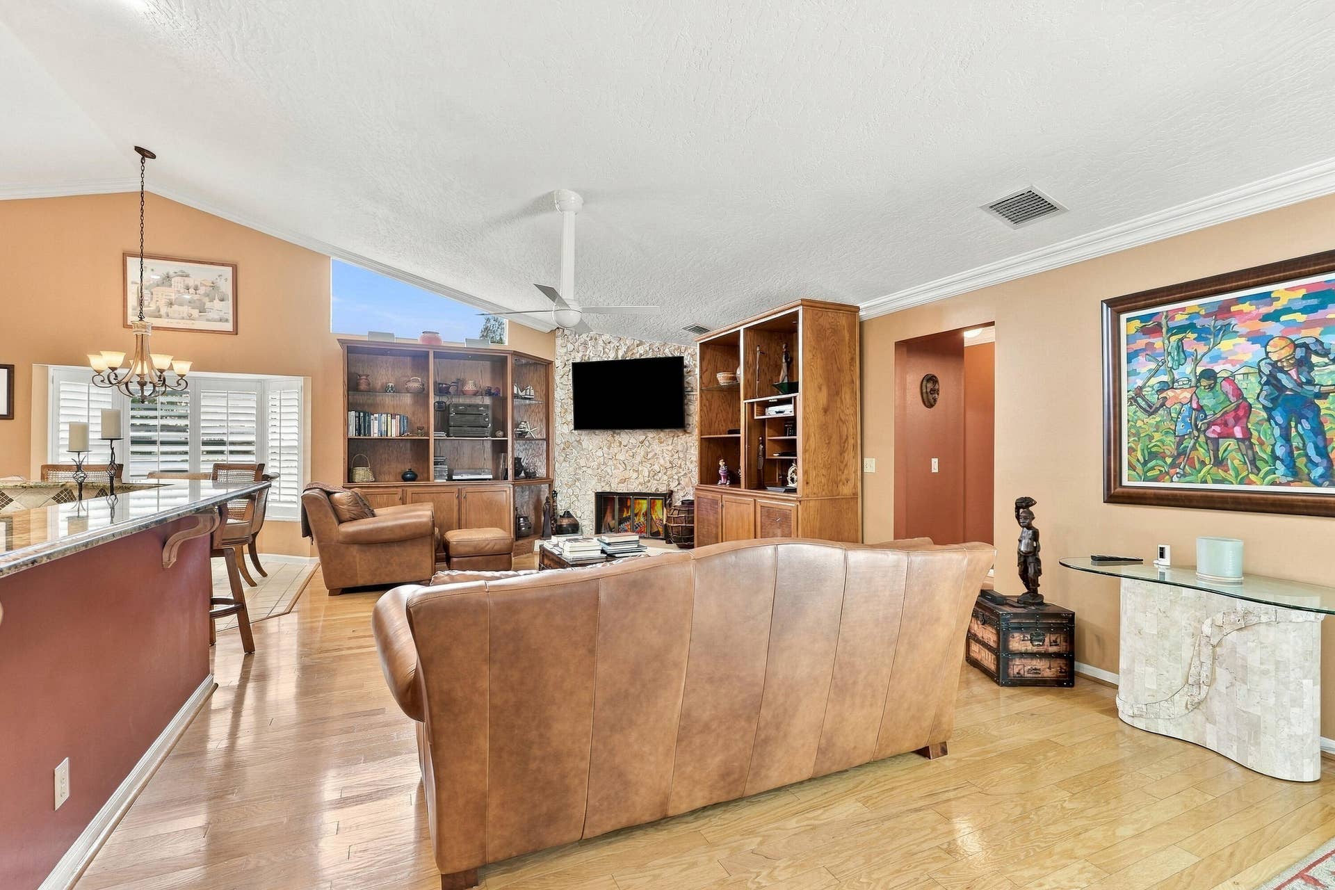 Vaulted ceilings with a ceiling fan, stone-clad fireplace, integrated wooden shelving and cabinetry, wood-finish flooring, and a breakfast bar with stone countertop