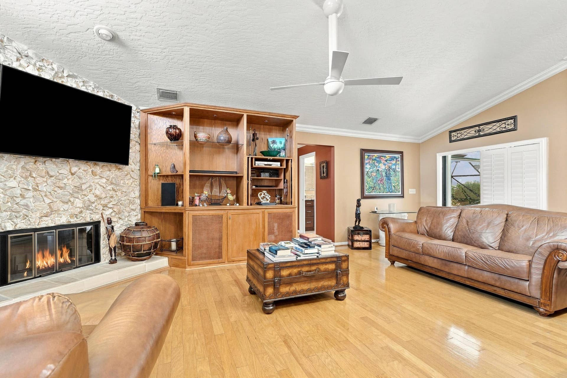 Vaulted ceiling with a ceiling fan, stone-clad fireplace, wood-finish flooring, built-in wooden shelving unit, and window with plantation shutters