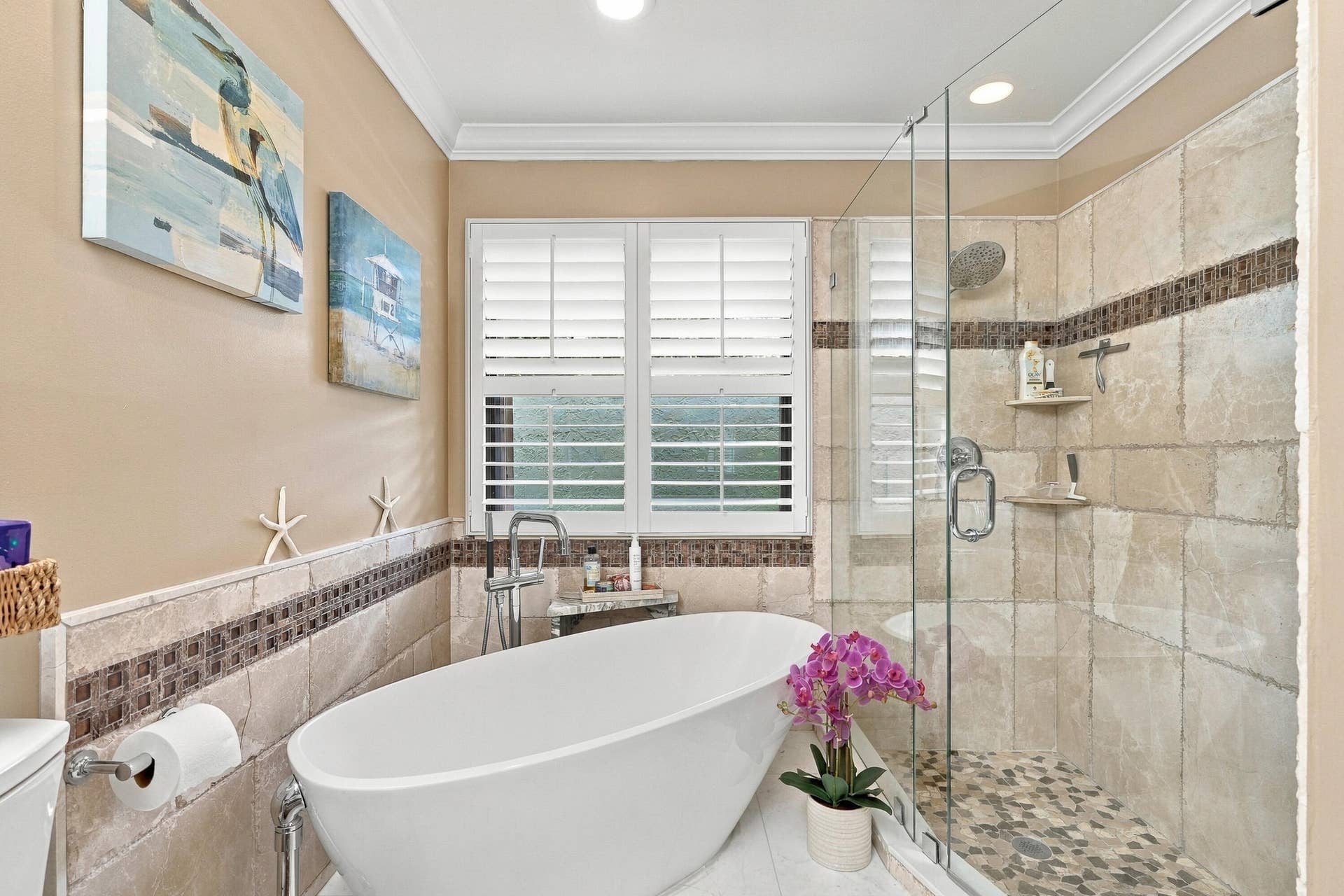 Freestanding soaking tub with floor-mounted faucet, walk-in shower with glass enclosure and pebble flooring, and window with plantation shutters