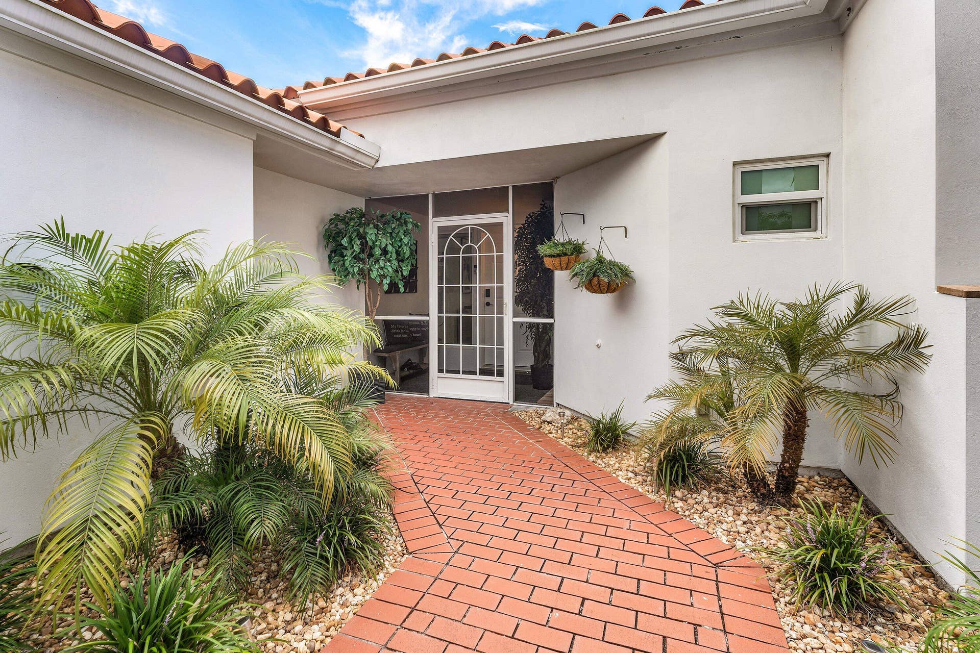 13138 Touchstone Place, Palm Beach Gardens, FL 33418 Entryway featuring a terra cotta tile roof, white stucco exterior, red brick pathway, screened arch-top door, and mature palm landscaping
