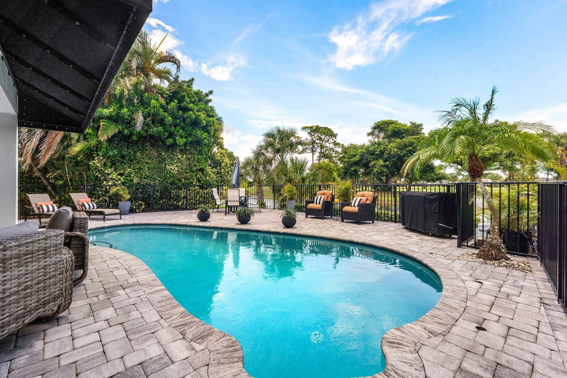 13138 Touchstone Place, Palm Beach Gardens, FL 33418 Curved swimming pool surrounded by paver decking, a black metal fence, and mature tropical landscaping