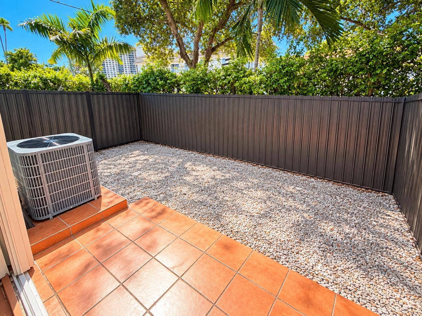 Tiled patio with gravel landscaping, surrounded by a privacy fence and lush foliage. Virtually staged.