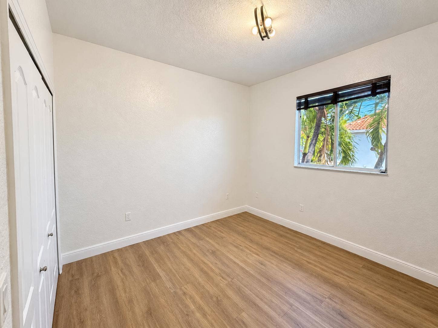 Wood-finish flooring with white baseboards, a window with horizontal blinds, and a built-in closet with bi-fold doors. Virtually staged.