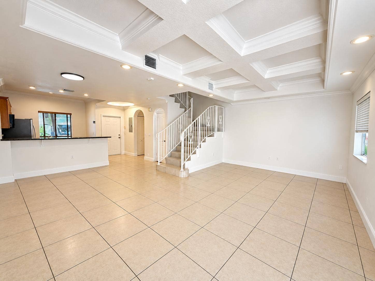 Spacious living area featuring large format tile flooring, coffered ceiling, recessed lighting, and a prominent staircase with a white railing. Virtually staged.