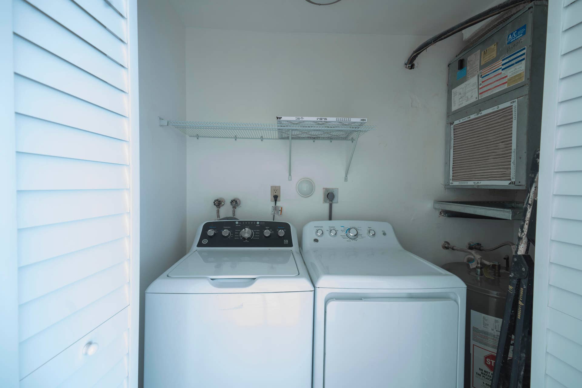 Louvered bifold doors concealing a laundry area featuring a white washer and dryer, wall-mounted wire shelving, and an air handling unit