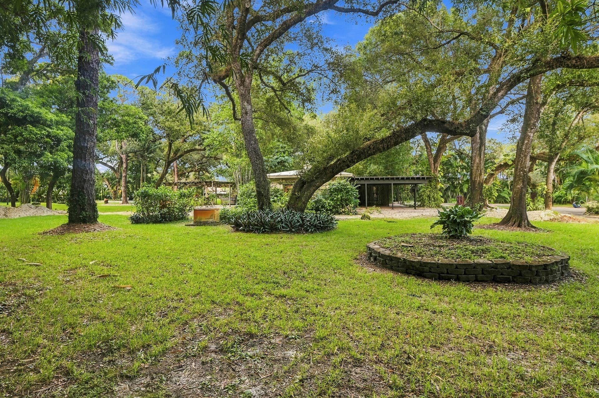 1549 SW 30th Place, Fort Lauderdale, FL 33315 Expansive lawn area with mature trees, a circular stone-edged planter, and a partially visible covered structure in the background
