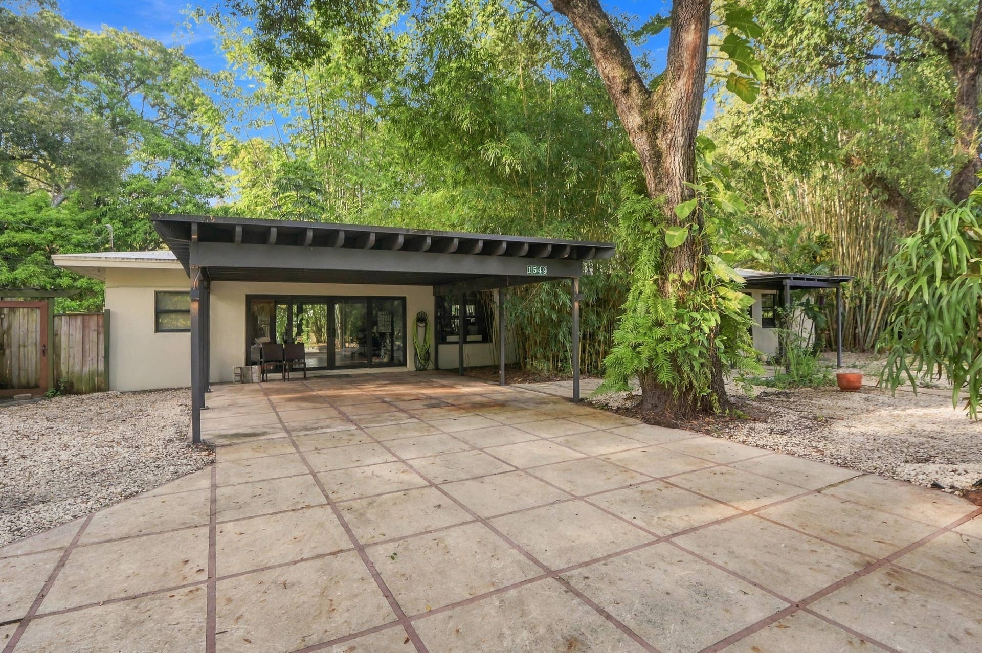 1549 SW 30th Place, Fort Lauderdale, FL 33315 Covered carport structure, expansive concrete driveway, natural landscaping with mature trees, and a beige exterior with dark-framed sliding glass doors