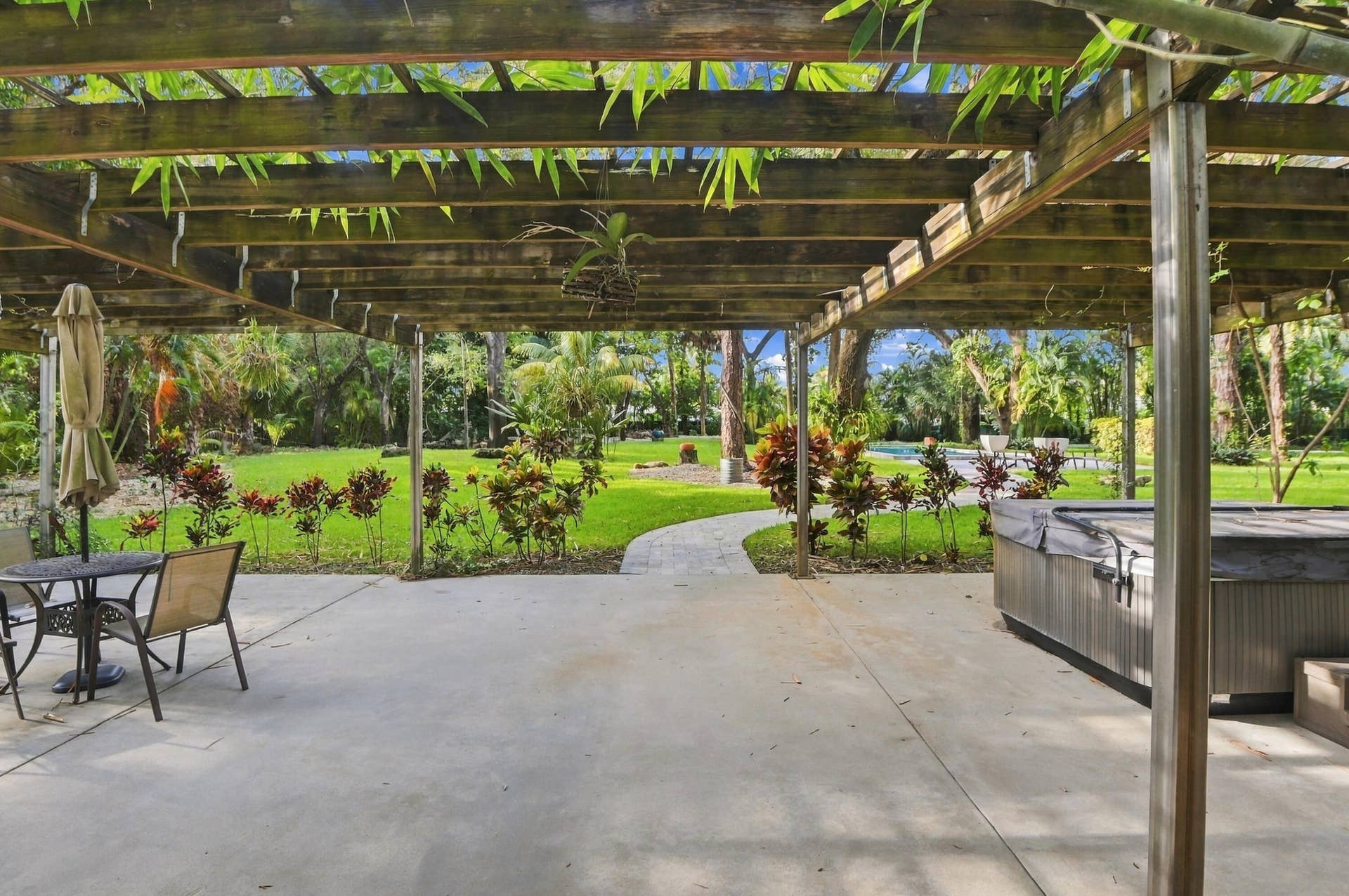 1549 SW 30th Place, Fort Lauderdale, FL 33315 Expansive concrete patio with a wooden pergola, leading to a manicured lawn with a winding stone pathway, lush tropical landscaping, and a built-in hot tub