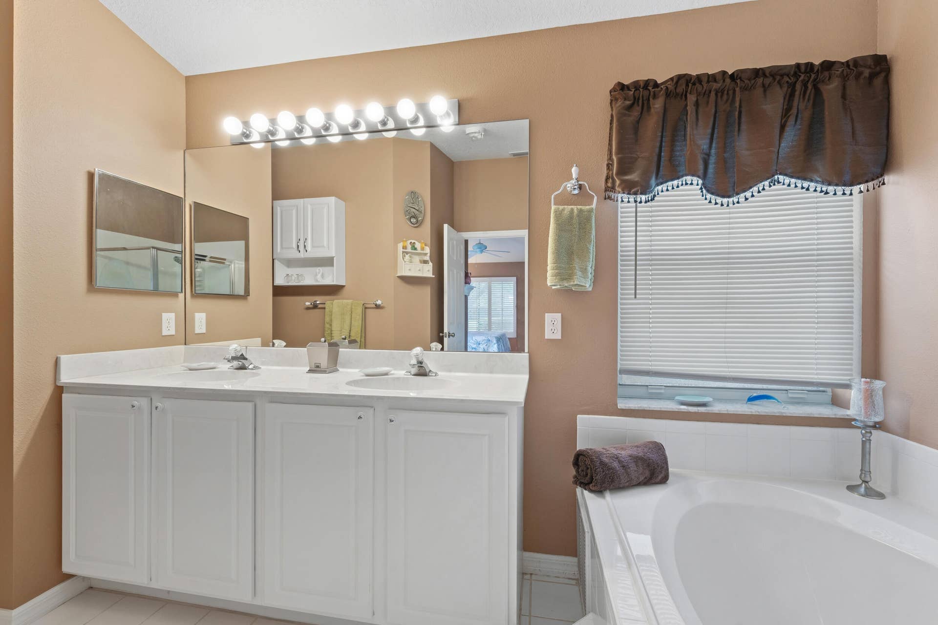 1649 SW ALVATON Avenue, Port St Lucie, FL 34953 Dual-sink vanity with white cabinetry and solid surface countertop, large wall-mounted mirror, integrated strip lighting, and soaking tub with white tile surround