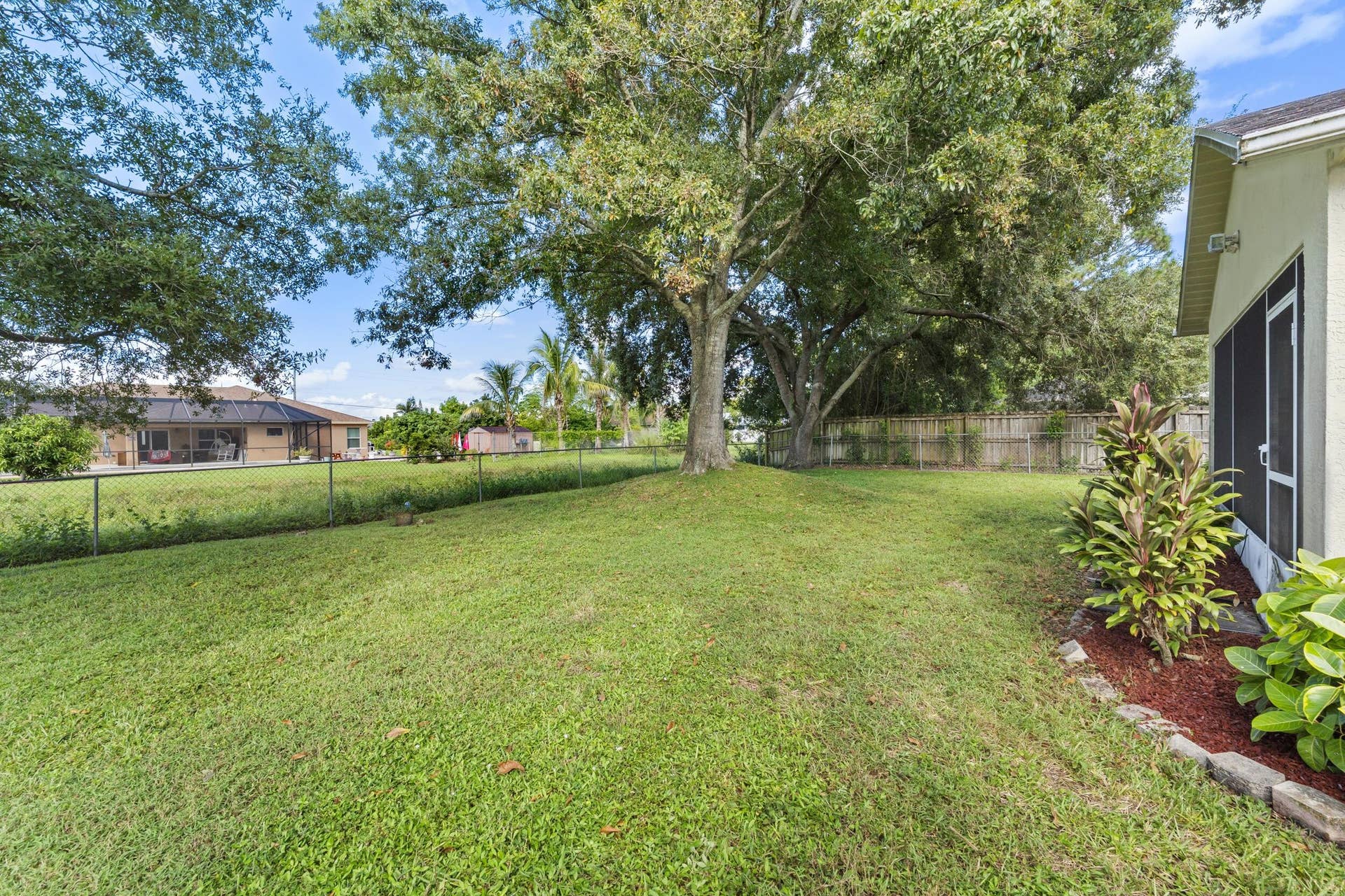 1649 SW ALVATON Avenue, Port St Lucie, FL 34953 Expansive backyard featuring a fenced perimeter, mature canopy trees, a grass lawn, and a stone-bordered garden bed