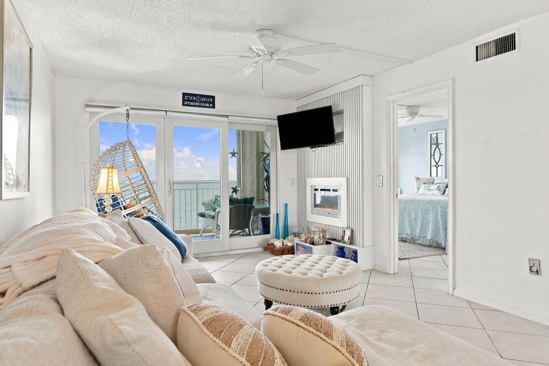 Bright, open living area featuring floor-to-ceiling glass doors and windows with ocean vistas, neutral tile flooring, ceiling fan, wall-mounted flat-screen television, and access to a furnished balcony overlooking the waterfront