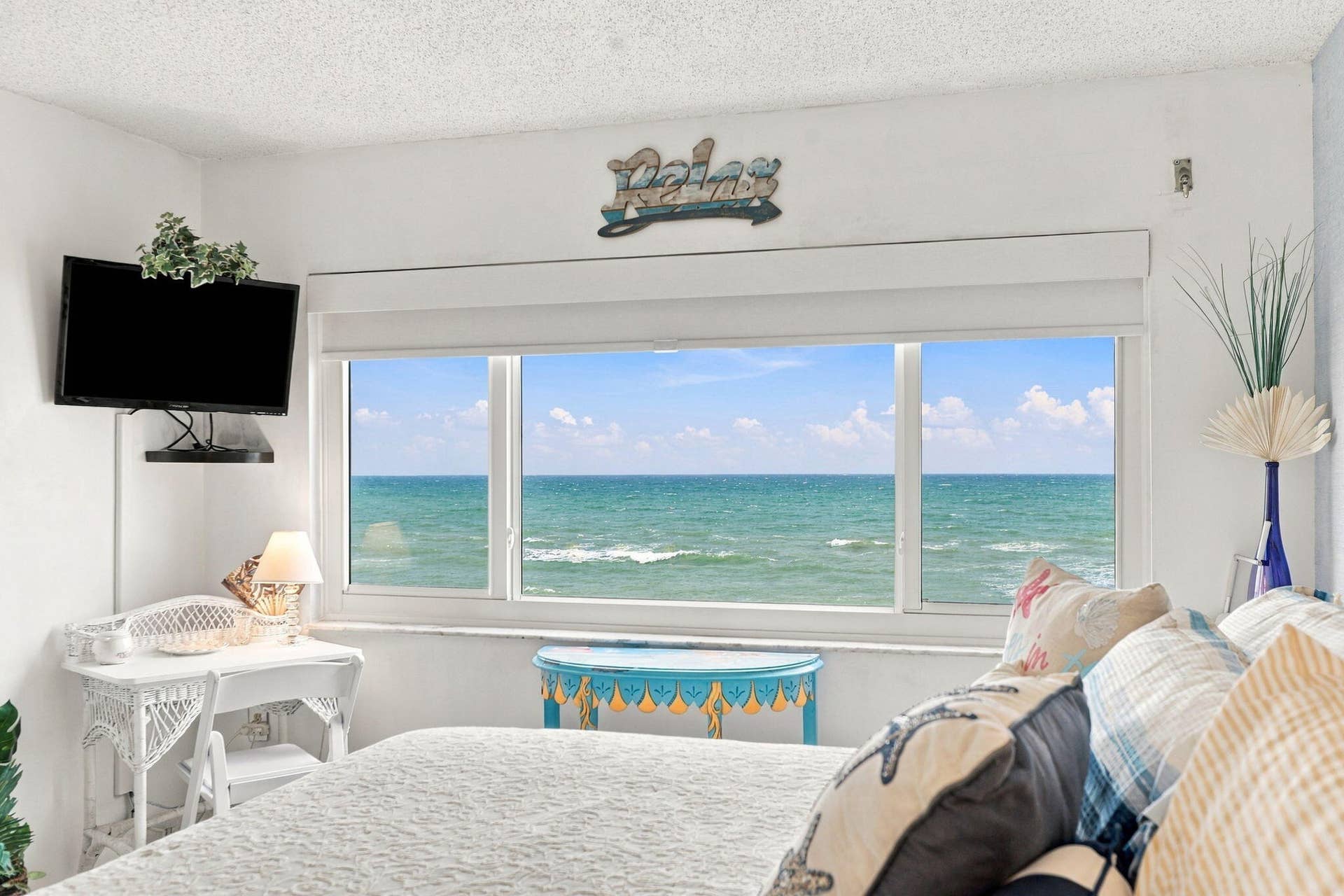 Oceanfront bedroom showcasing expansive floor-to-ceiling windows framing direct water vistas, white wicker furnishings, neutral bedding, and a mounted flat-screen television