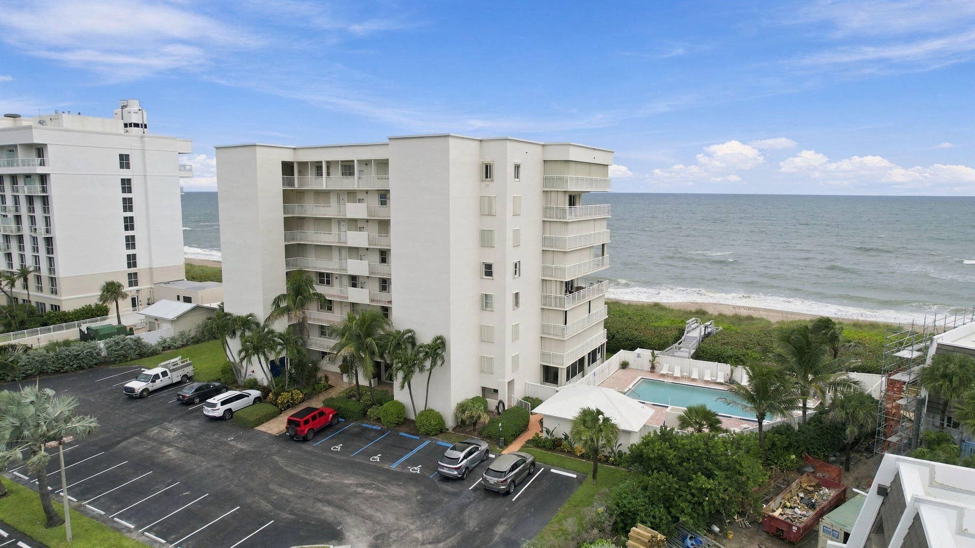 Coastal condominium building featuring multiple private balconies, a swimming pool with a surrounding deck, and direct beach access via a boardwalk