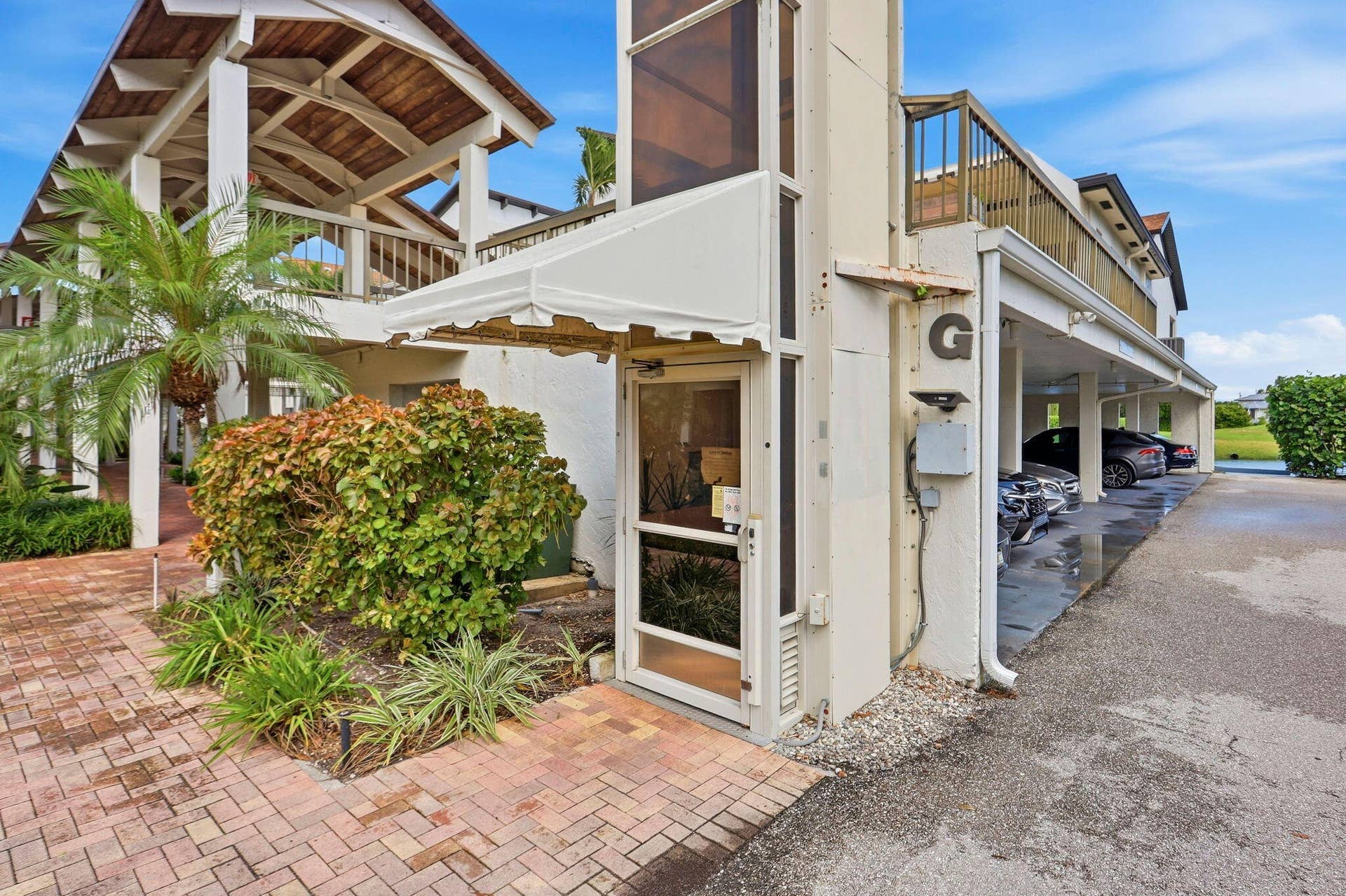 900 Greensward Lane, #209-G, Delray Beach, FL 33445 Outdoor brick paver walkway leading to an entry with a white awning, flanked by mature landscaping and a palm tree