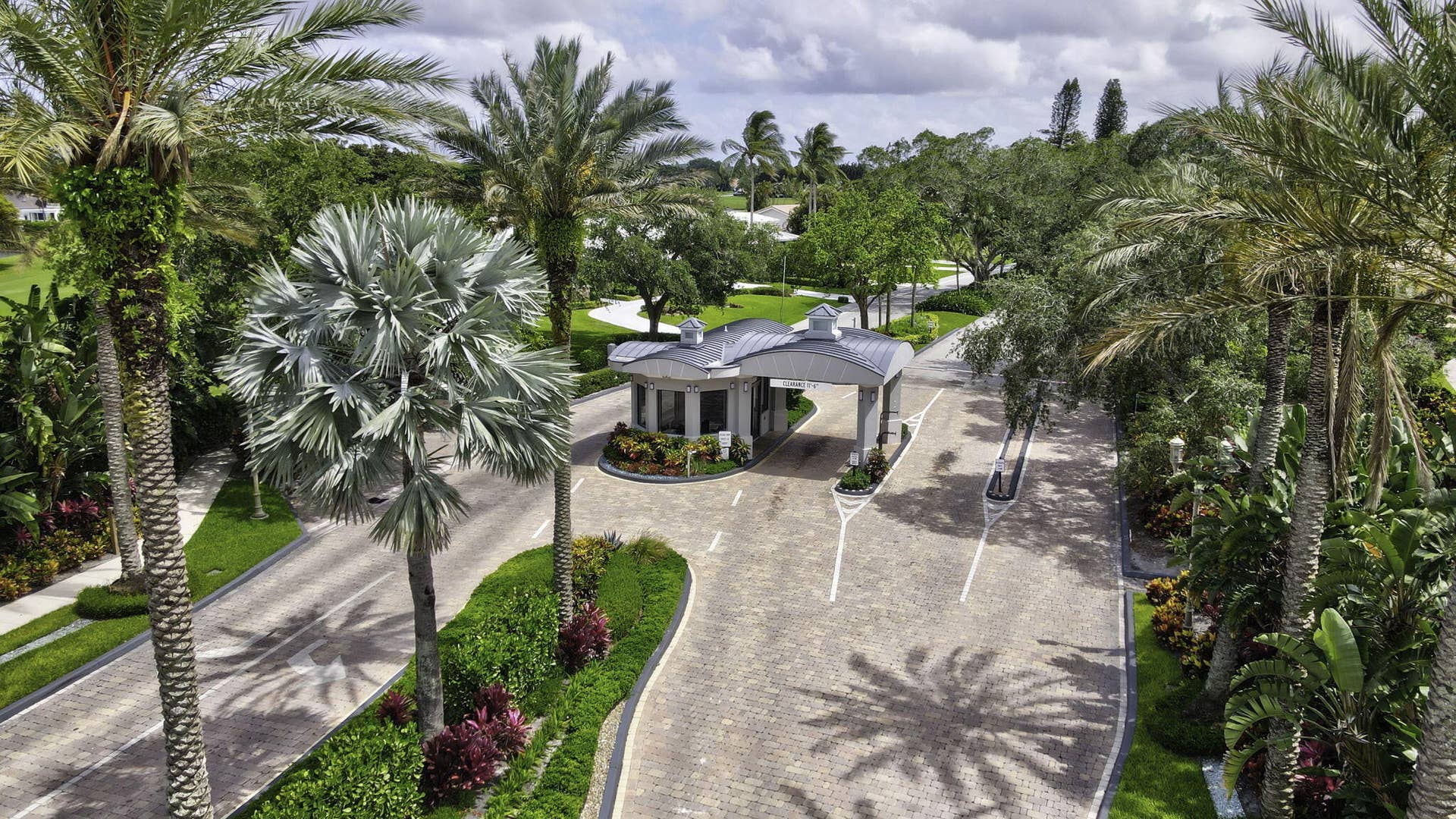 900 Greensward Lane, #209-G, Delray Beach, FL 33445 Gated entry with a stone-finish driveway, mature palm trees, and lush landscaping