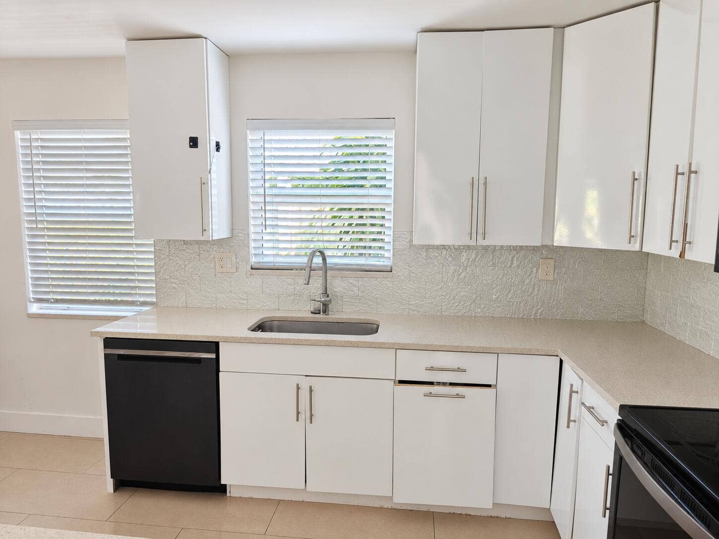 White cabinetry with brushed nickel hardware, light-toned countertops, a stainless steel sink with a gooseneck faucet, a textured backsplash, and a black dishwasher
