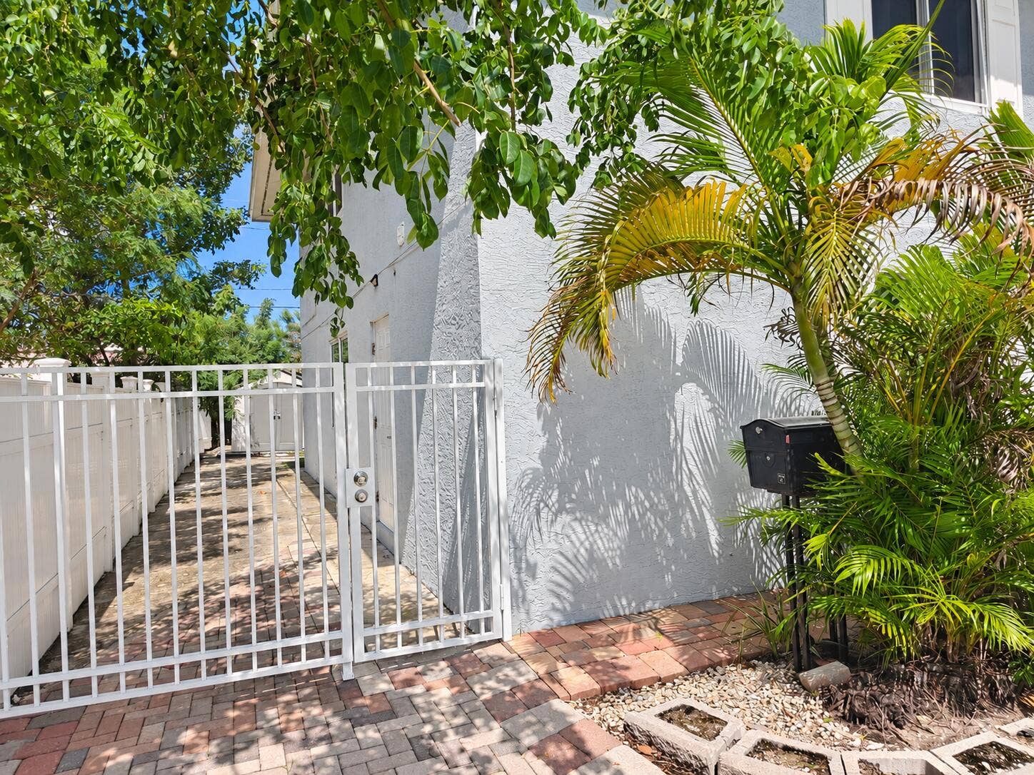White stucco exterior, white metal gate, brick paver walkway, mature palm trees, lush landscaping