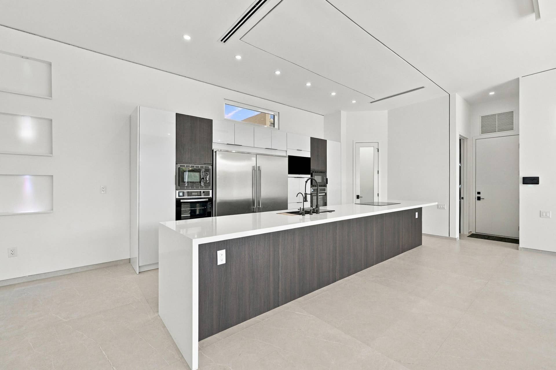 Modern kitchen featuring a large waterfall-edge island with wood-finish cabinetry, stainless steel appliances including a built-in refrigerator and wall-mounted oven, white quartz countertops, and polished concrete flooring throughout