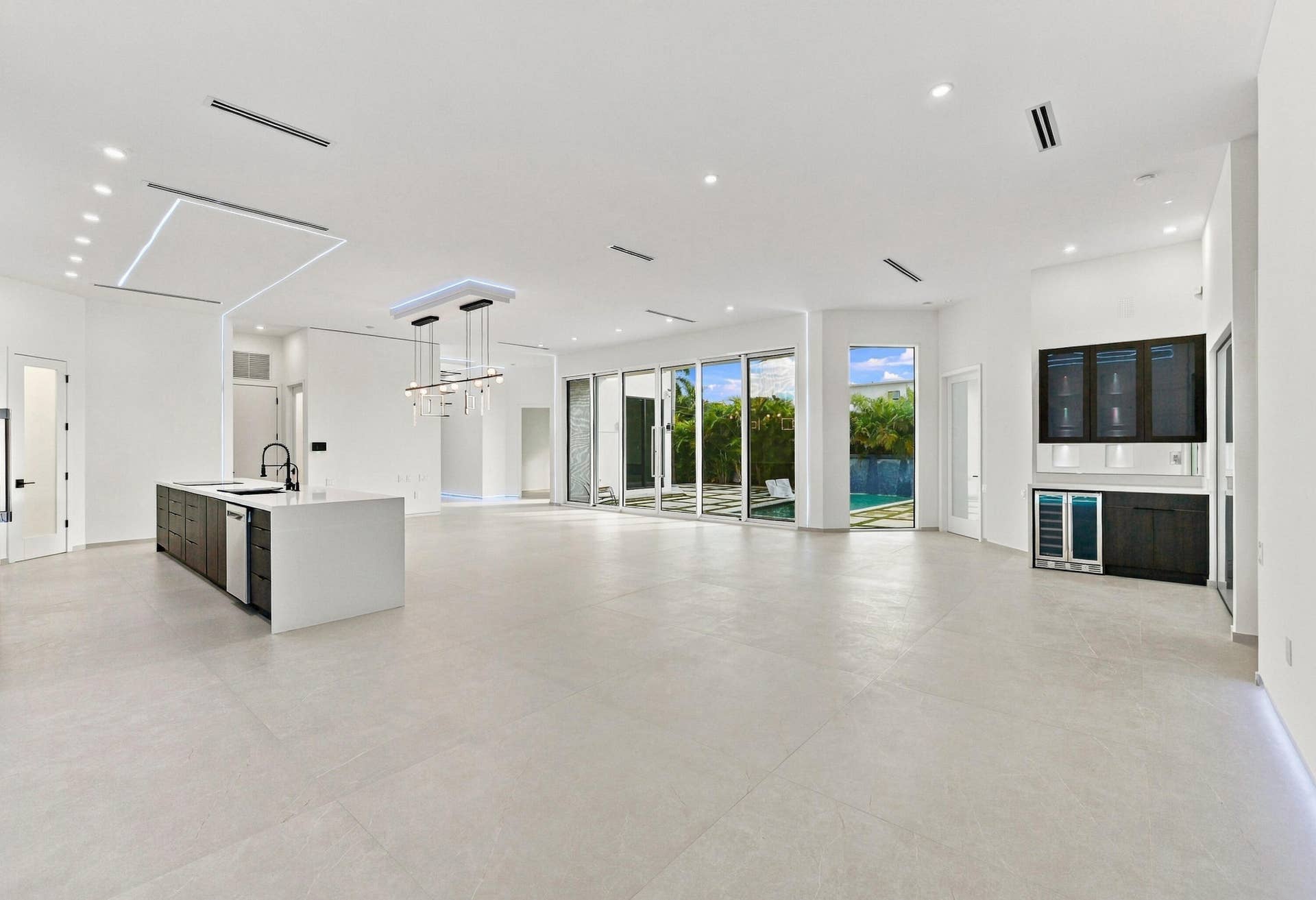 Expansive open-concept living space featuring polished light-toned flooring throughout, abundant natural light from multiple glass sliding doors and windows, a modern kitchen with waterfall-edge island and stainless steel appliances, recessed and linear ceiling lighting, and built-in entertainment cabinetry with integrated display screens