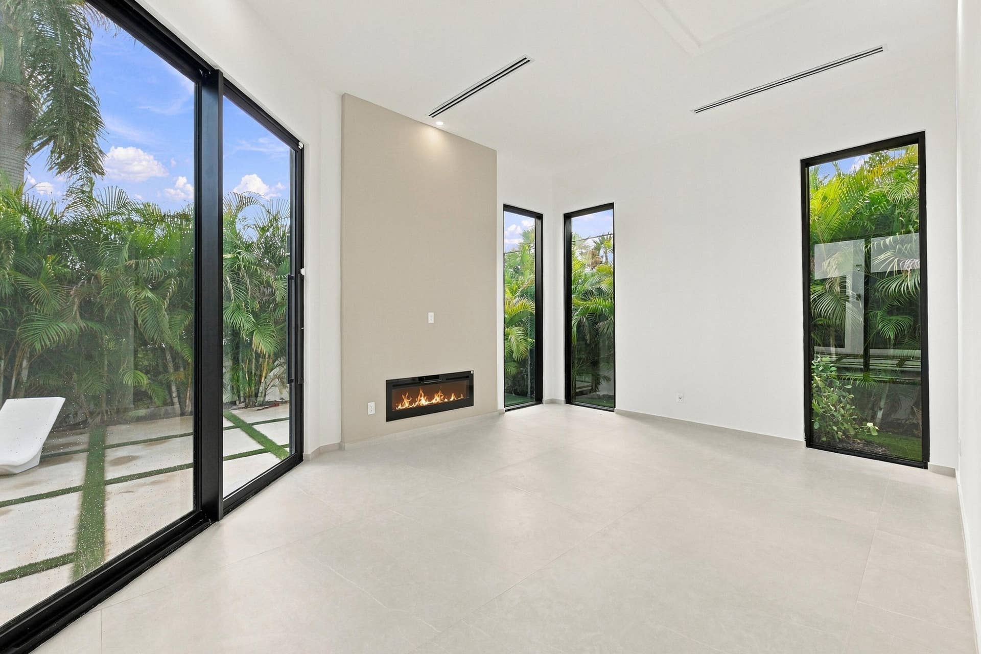 Bright, open interior featuring polished concrete flooring, floor-to-ceiling glass sliding doors with black frames, linear fireplace with stone surround, recessed ceiling lighting, and expansive windows framing mature landscaping and outdoor grounds