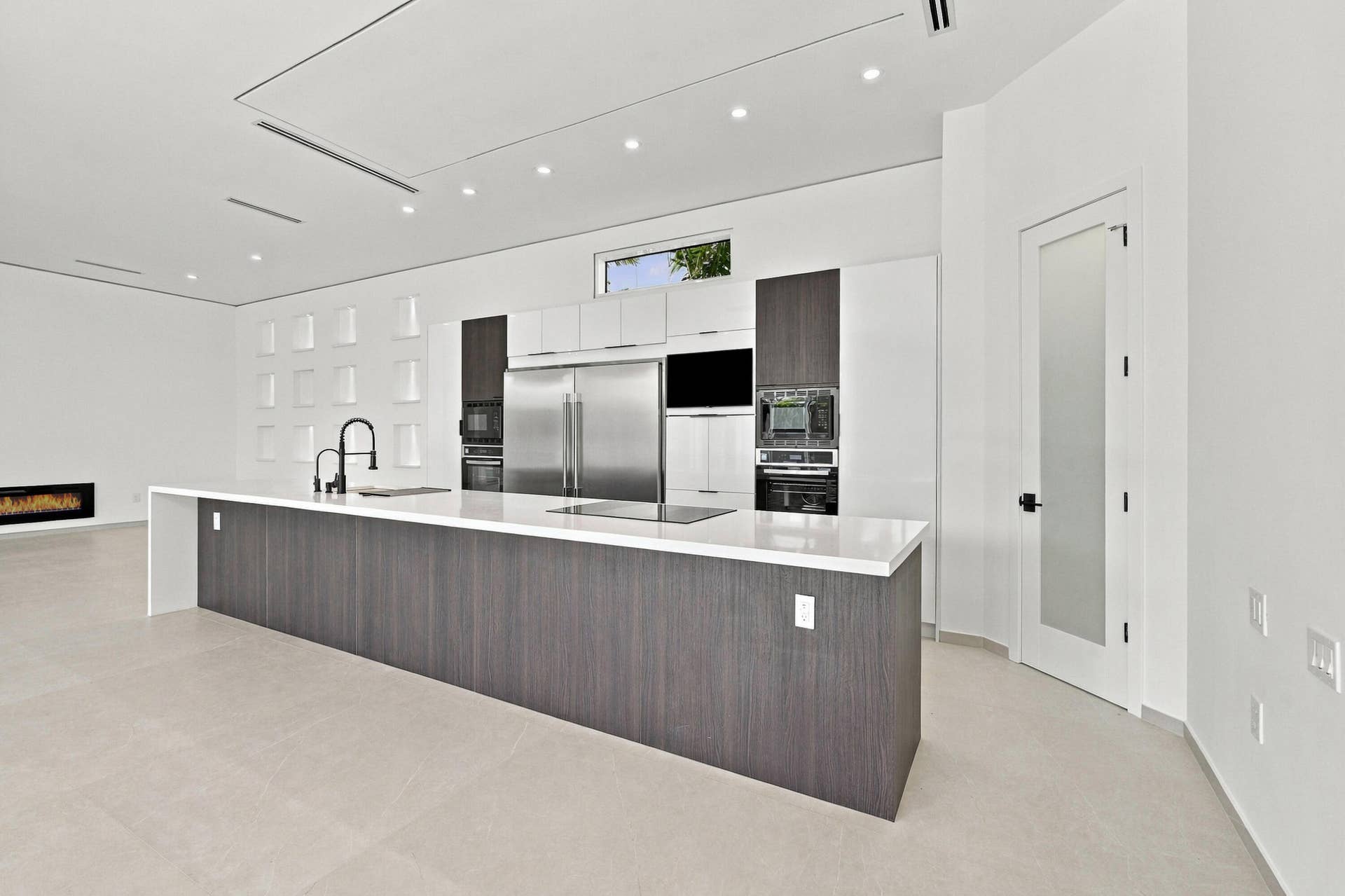 Contemporary kitchen featuring a large waterfall-edge island with dark wood-finish cabinetry, stainless steel appliances including a built-in refrigerator and wall-mounted oven, white quartz countertops, and modern pendant lighting