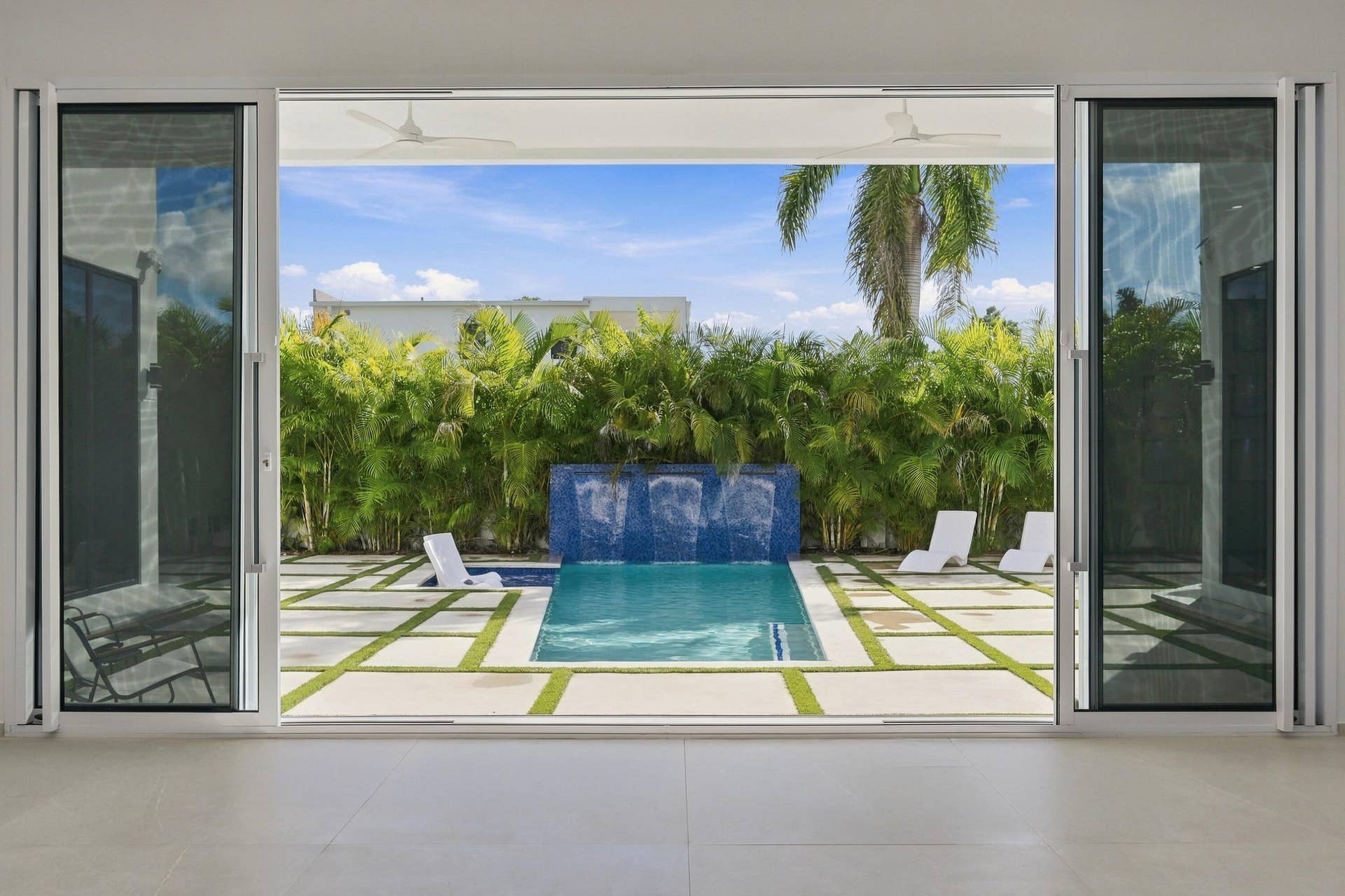 Seamless indoor-outdoor living with floor-to-ceiling glass doors opening to a resort-style backyard featuring a geometric paved terrace, resort pool with integrated water feature wall, mature landscaping with specimen palms and trees, and lounge seating areas