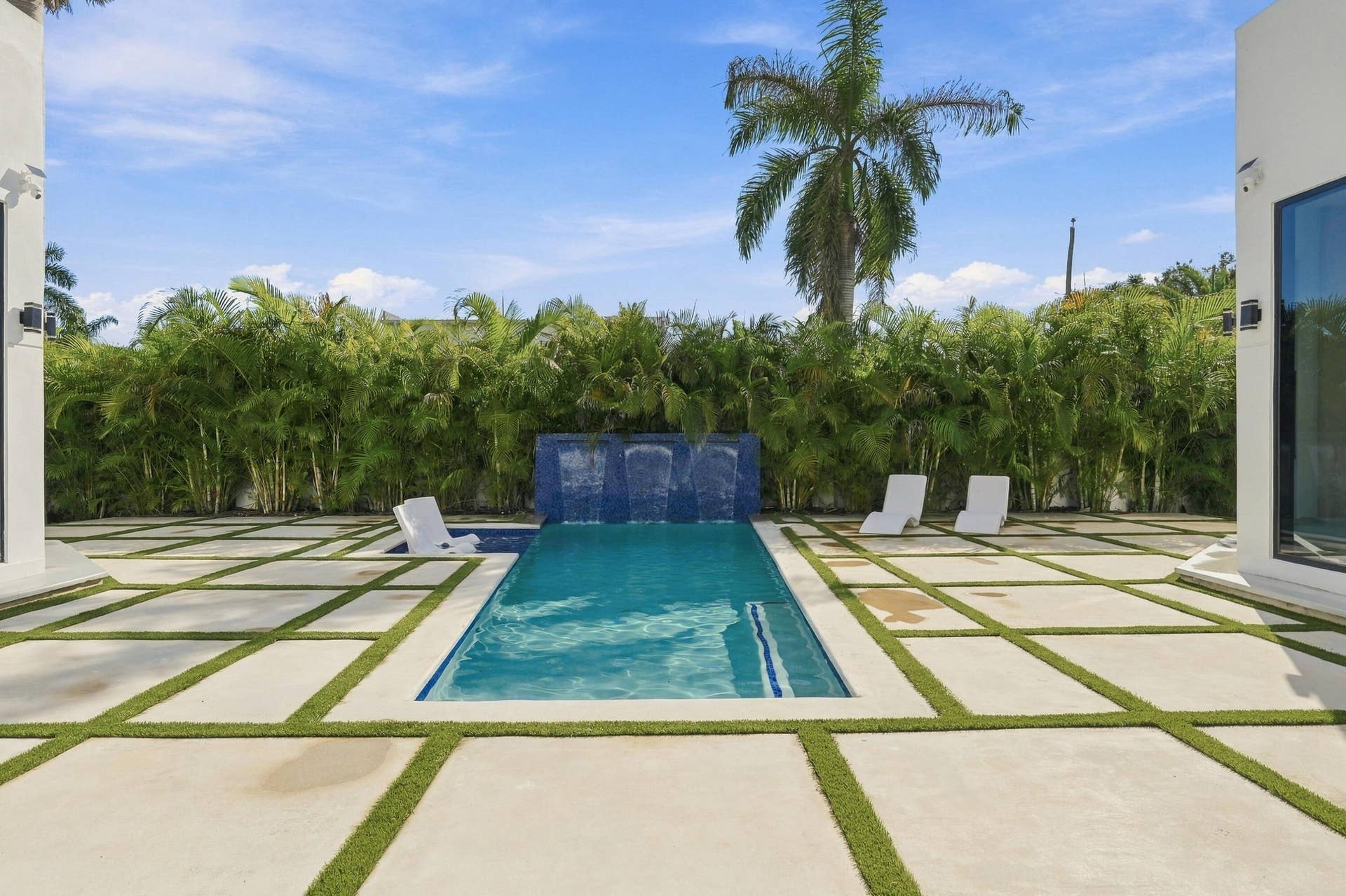 Rectangular swimming pool with a tile waterfall feature, surrounded by paver patio with artificial grass accents, and a lush privacy hedge