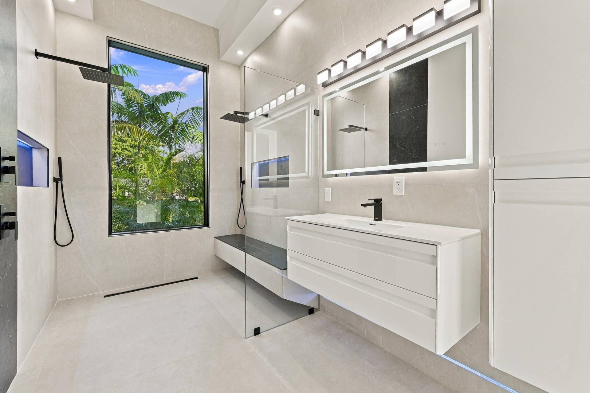 Spa-inspired primary bathroom featuring a frameless glass-enclosed shower with rainfall showerhead and handheld fixture, floating vanity with integrated sink and matte black hardware, large illuminated mirror with dual sconces, light-finish tile flooring, and abundant natural light from an oversized window