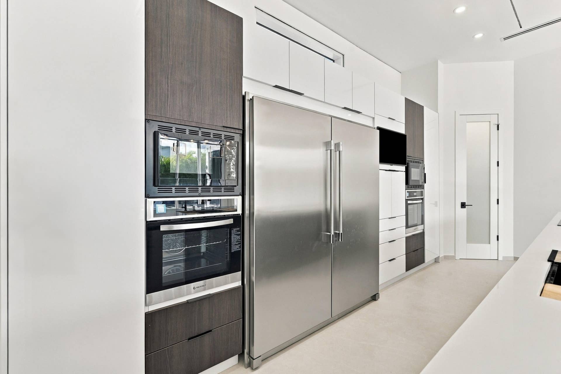 Contemporary kitchen featuring stainless steel side-by-side refrigerator with dual doors, built-in wall ovens with black glass fronts, integrated microwave, white cabinetry with wood-finish accents, and light wood-finish flooring throughout the open layout