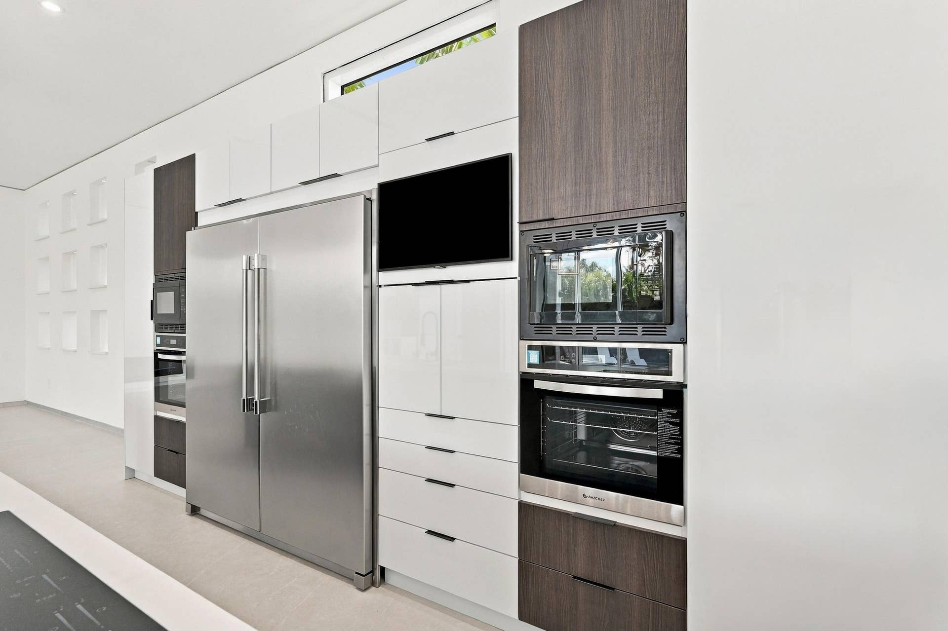 Contemporary kitchen featuring stainless steel dual-door refrigerator, integrated wall-mounted oven and microwave, flat-panel cabinetry in white and wood-finish finishes, quartz countertops, and built-in appliances