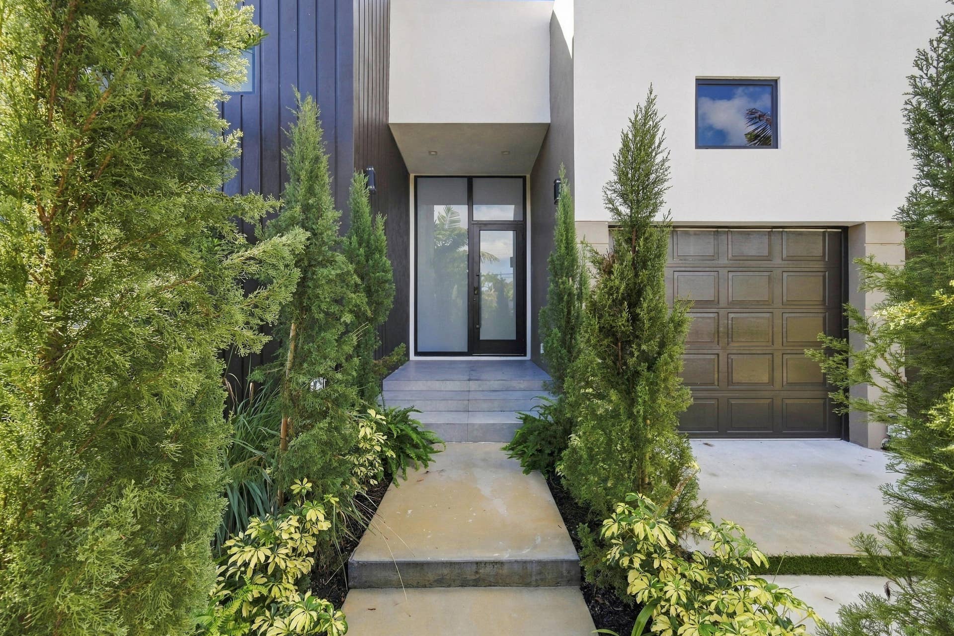 Modern architectural entry featuring a linear stone-paved walkway flanked by columnar evergreen landscaping, leading to a minimalist facade with clean lines, dark-framed glass entry doors, bronze panel garage door, and a small window accent detail on the upper facade