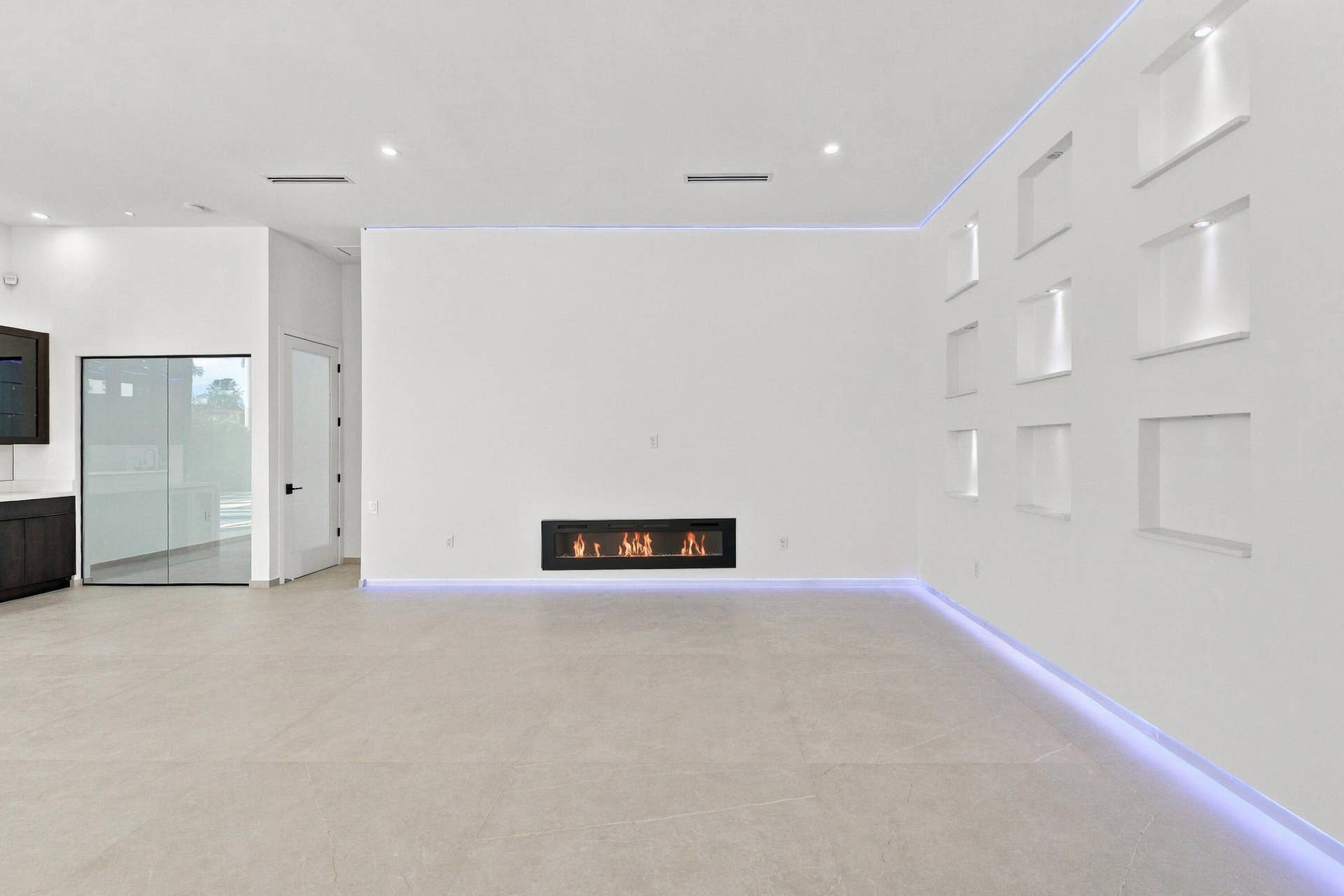 Sophisticated open-concept living space featuring light-toned polished concrete flooring, expansive white walls with architectural detail panels, recessed ceiling lighting with integrated LED accent strips, and a contemporary linear gas fireplace with glass surround