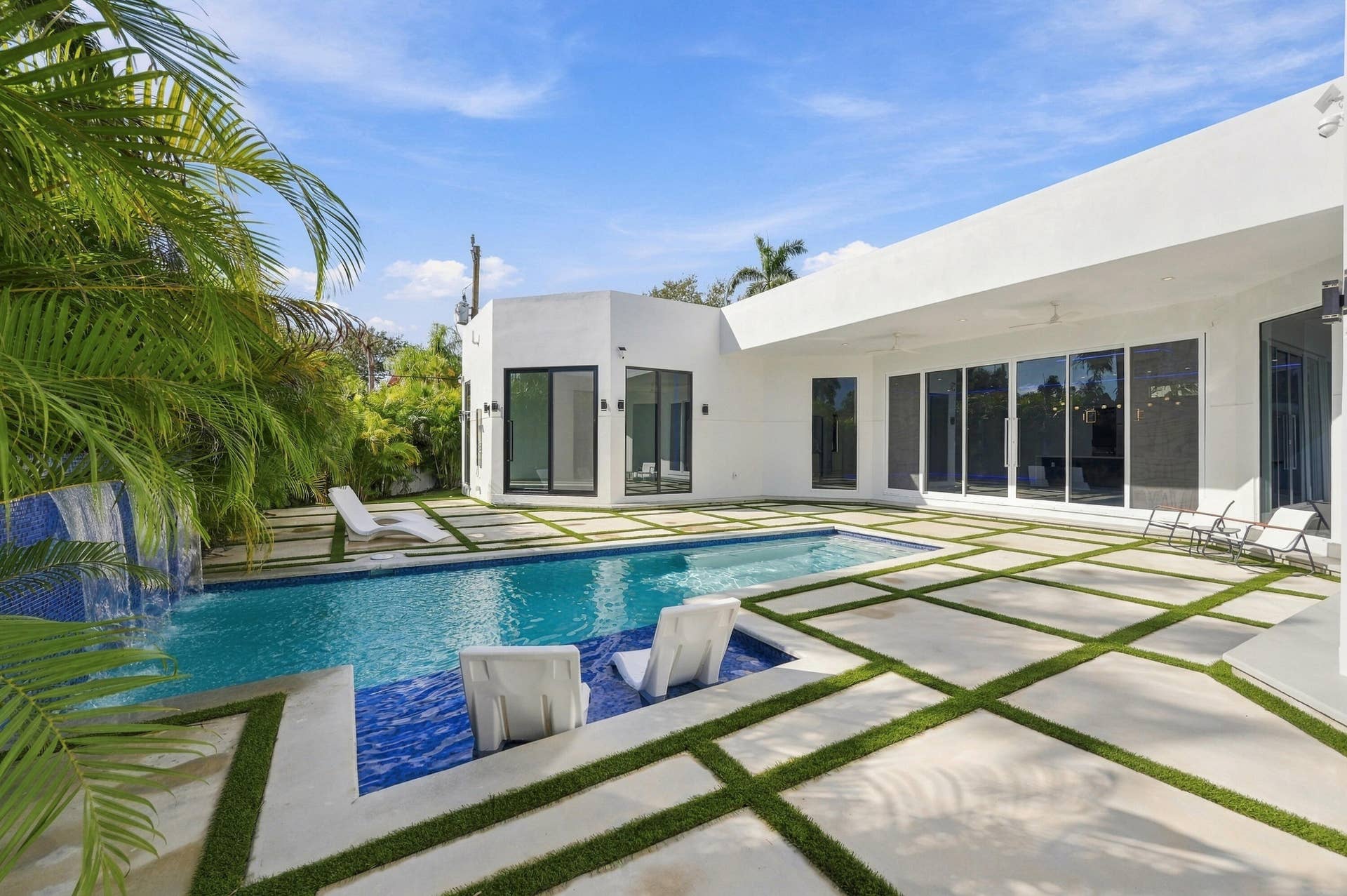 Modern residence featuring a resort-style swimming pool with crystal-clear waters, expansive hardscape patio with geometric inset planting beds, mature tropical landscaping including palm trees, clean-lined white exterior with floor-to-ceiling windows, and lounge seating areas overlooking the pool deck