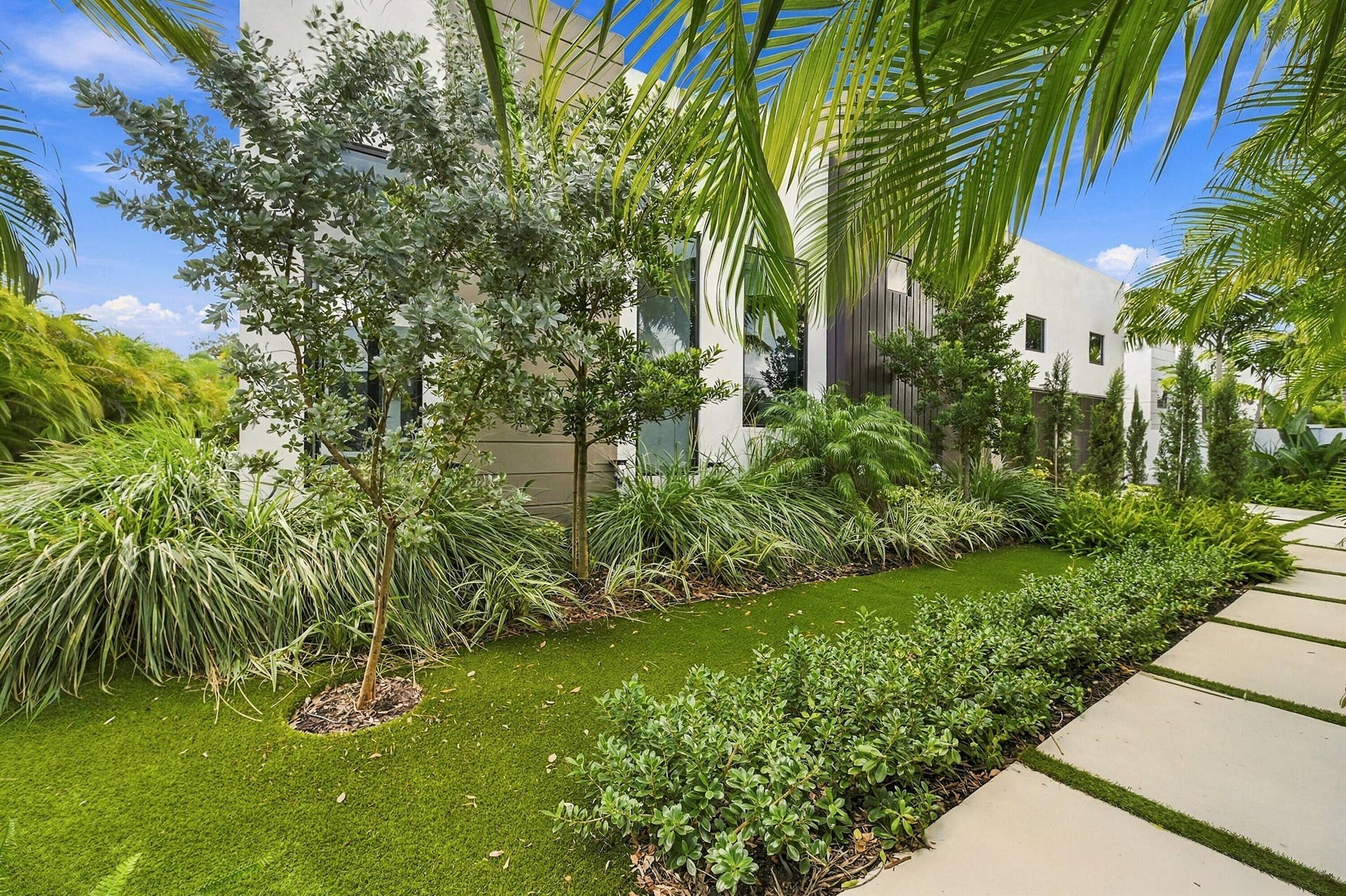 Lush landscaped grounds featuring mature palms and tropical plantings, manicured lawn, and linear concrete pathway leading to the residence