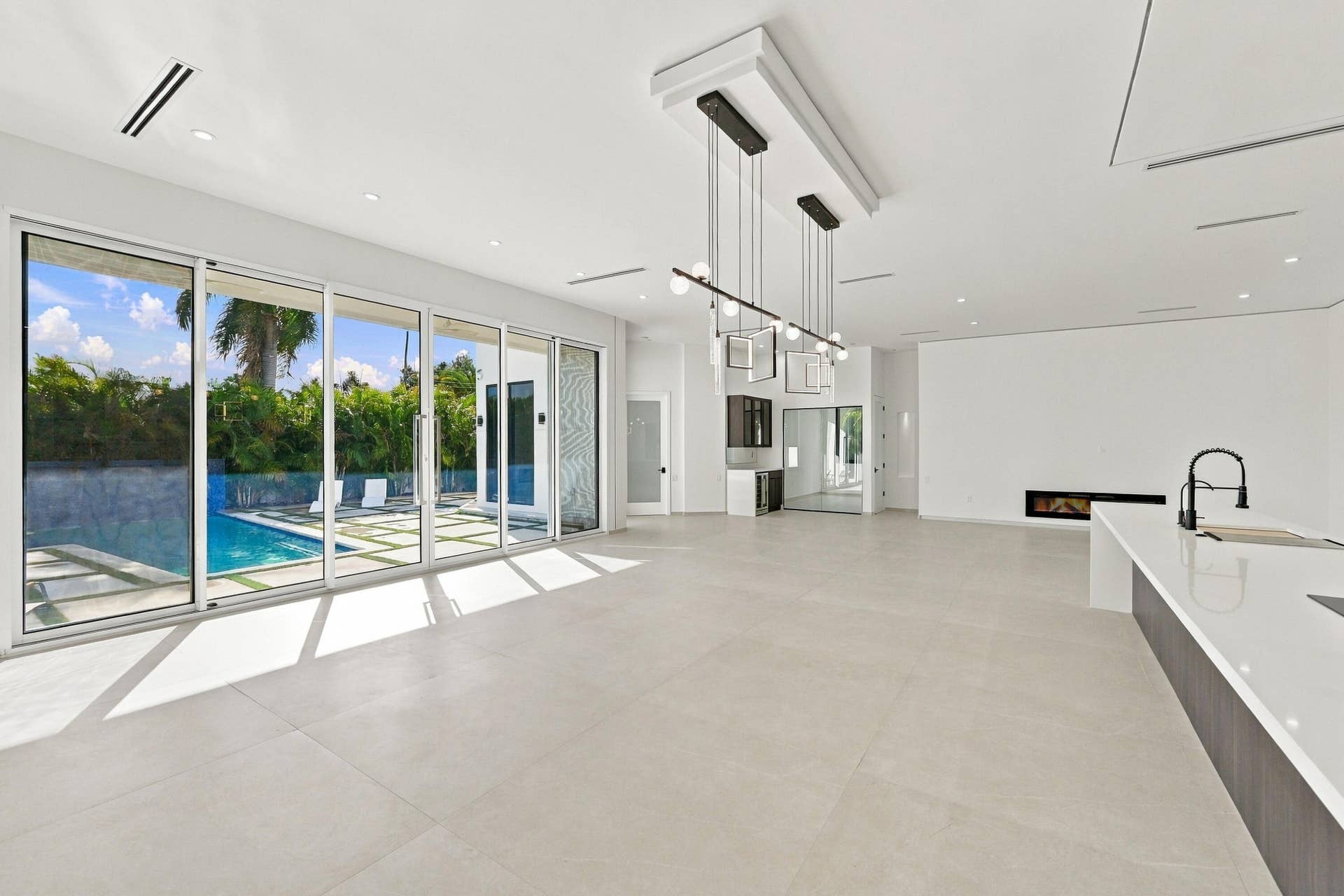 Sophisticated open-concept living space featuring polished light-toned stone flooring, floor-to-ceiling multi-panel glass doors and windows overlooking the pool and landscaped grounds, modern linear pendant lighting, and a contemporary kitchen with white cabinetry and stainless steel appliances