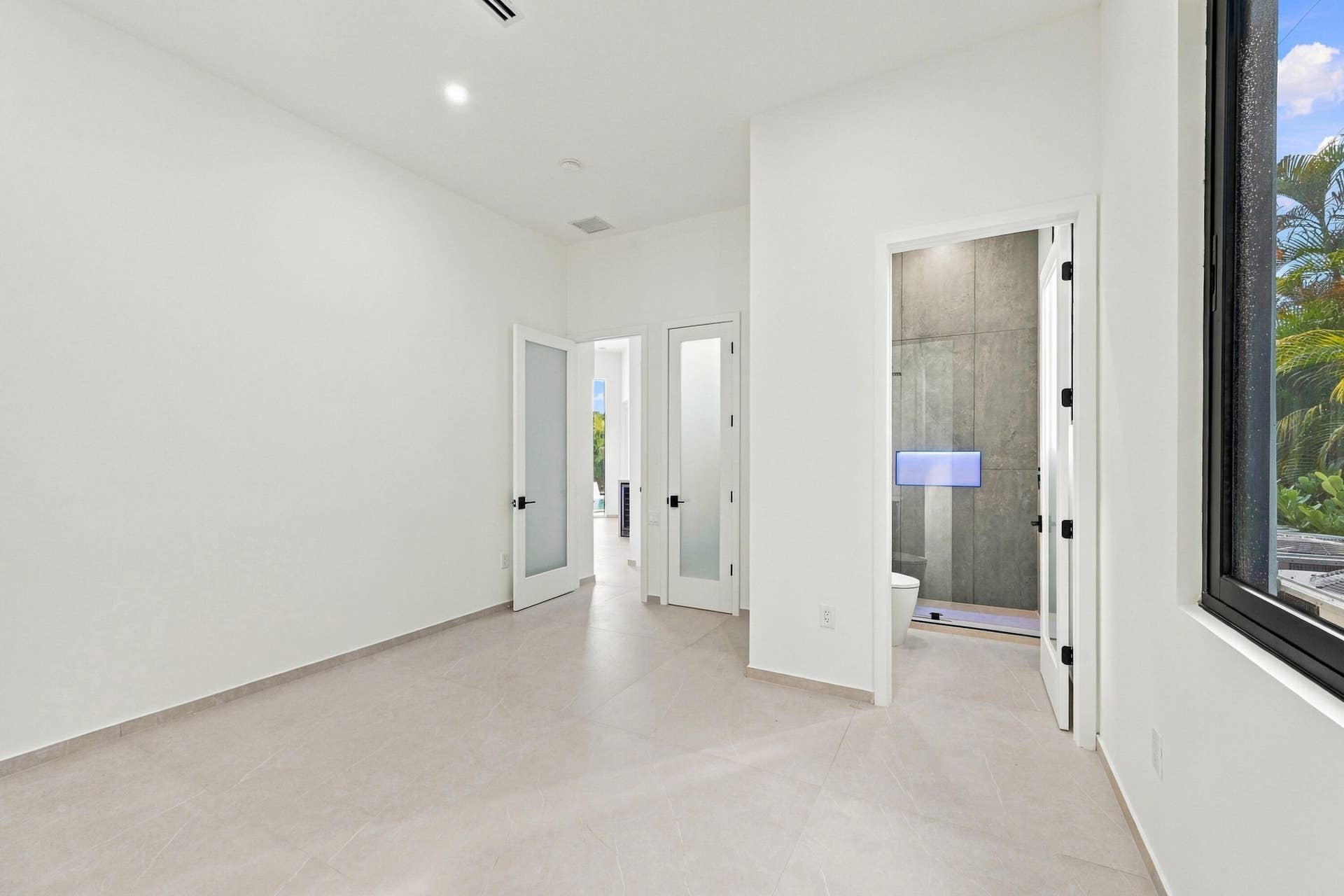 Bright, open primary suite featuring polished concrete flooring, multiple glass-panel French doors with black hardware, recessed ceiling lighting, and abundant natural light from oversized windows