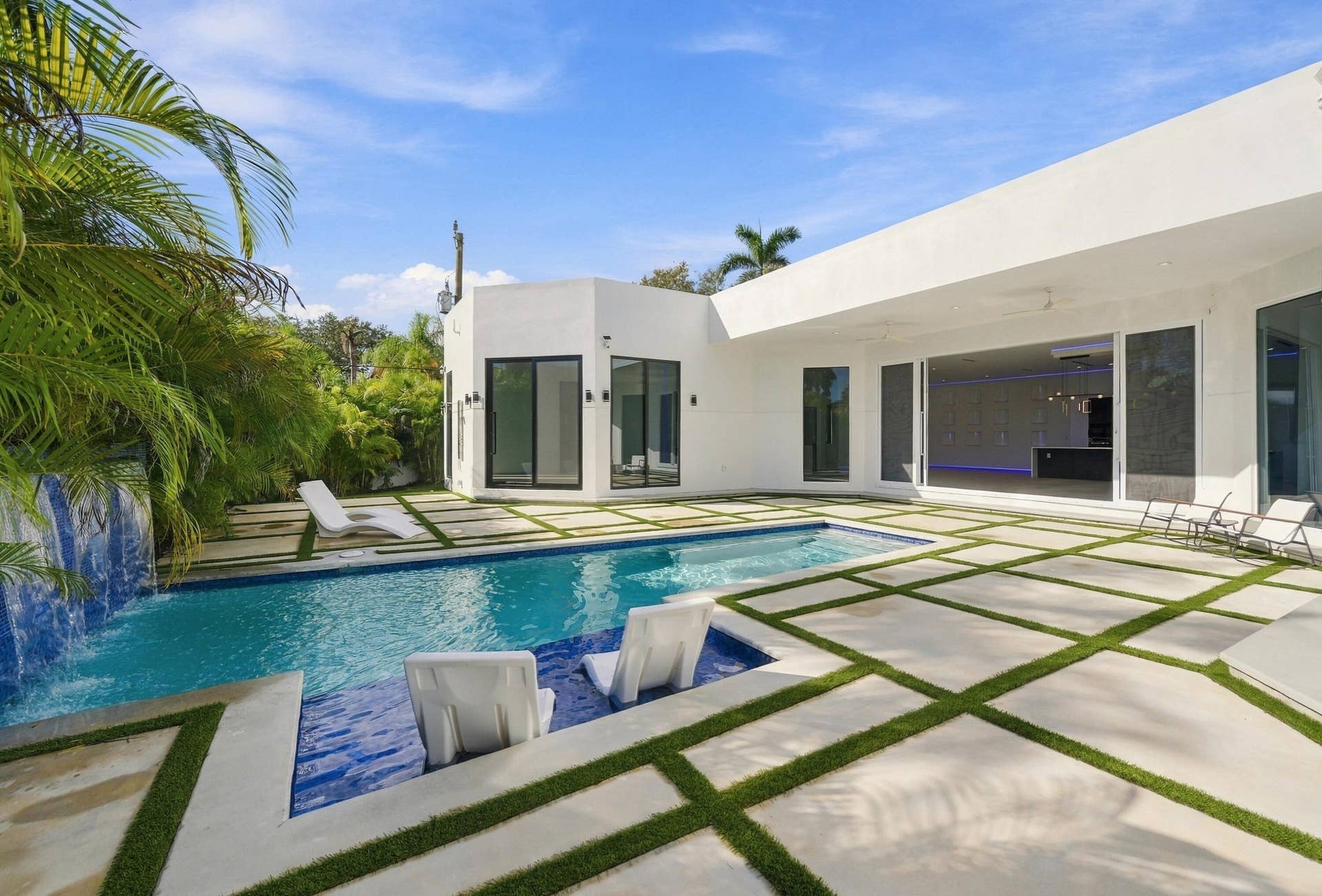 Modern poolside oasis featuring a resort-style saltwater pool with integrated spa seating, geometric concrete decking with contrasting grass inlays, and a clean-lined white stucco residence with floor-to-ceiling glass doors and operable windows