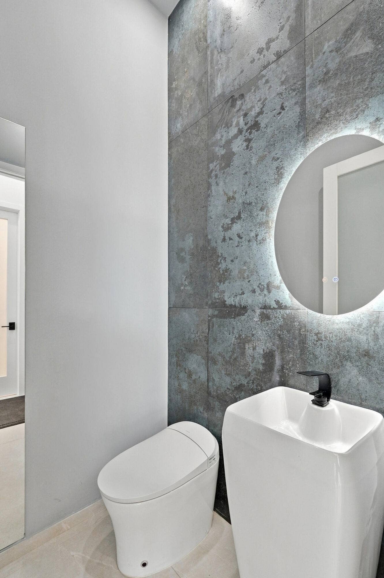 Modern bathroom featuring a sleek vessel sink with matte black wall-mounted faucet, illuminated circular mirror with integrated lighting, and a contemporary toilet