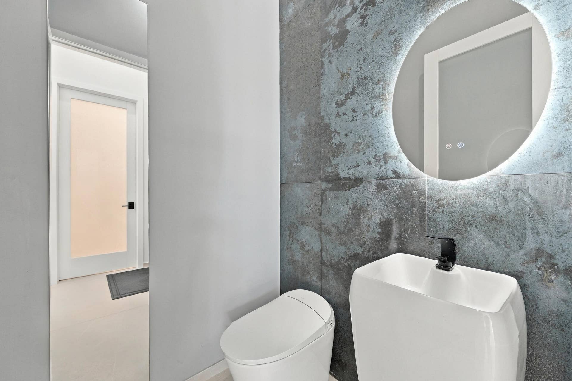 Modern bathroom featuring a sleek ceramic vessel sink with matte black faucet, illuminated circular mirror with integrated LED lighting, and polished white cabinetry